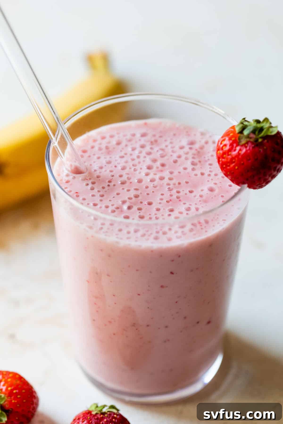 Another appealing shot of a strawberry banana smoothie in a glass with a straw and a fresh strawberry on the rim, emphasizing its refreshing quality.