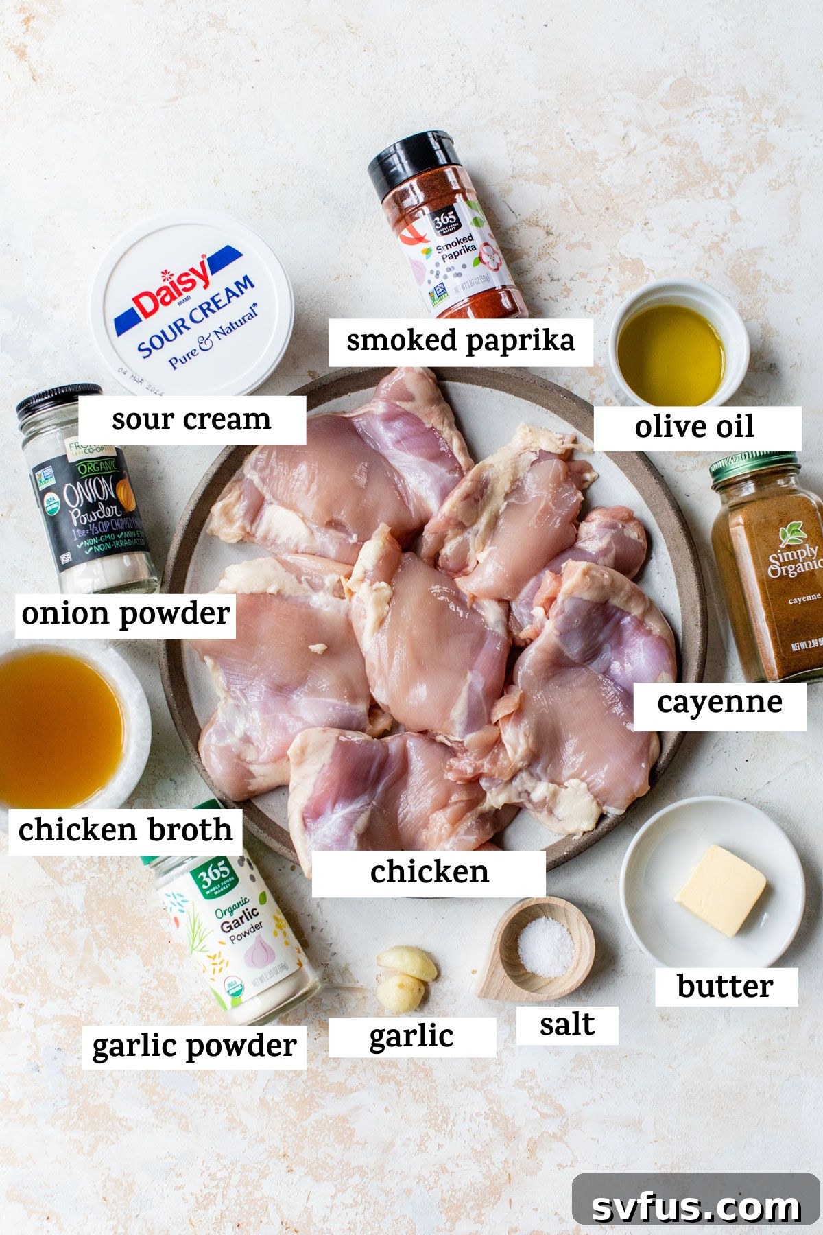 All ingredients for paprika chicken laid out on a wooden table, including chicken thighs, smoked paprika, garlic, onion powder, salt, cayenne, olive oil, butter, chicken broth, and sour cream.