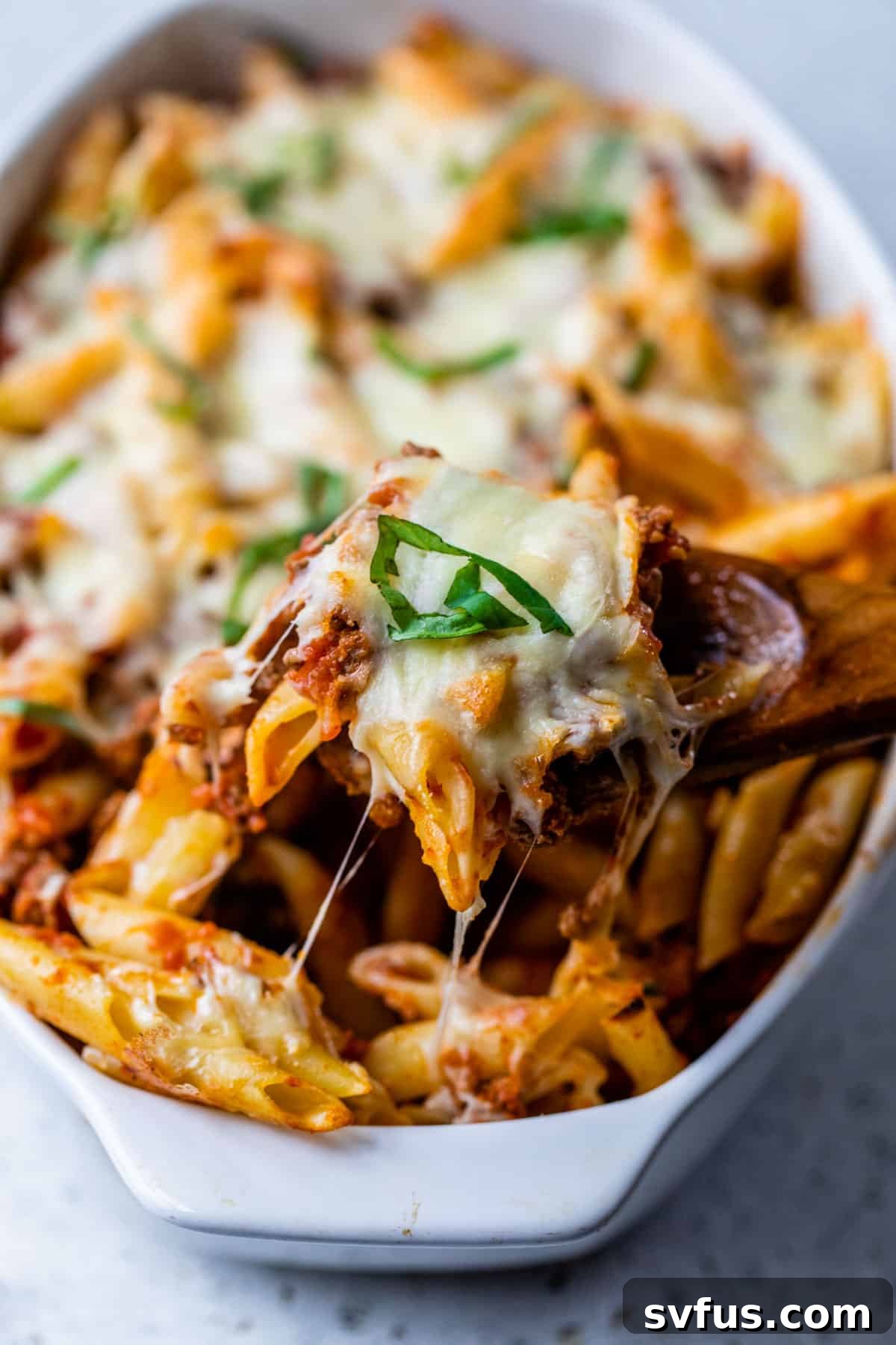 A spoonful of vegan baked ziti being scooped out of a large casserole dish, showing layers of pasta, sauce, and melted cheese.