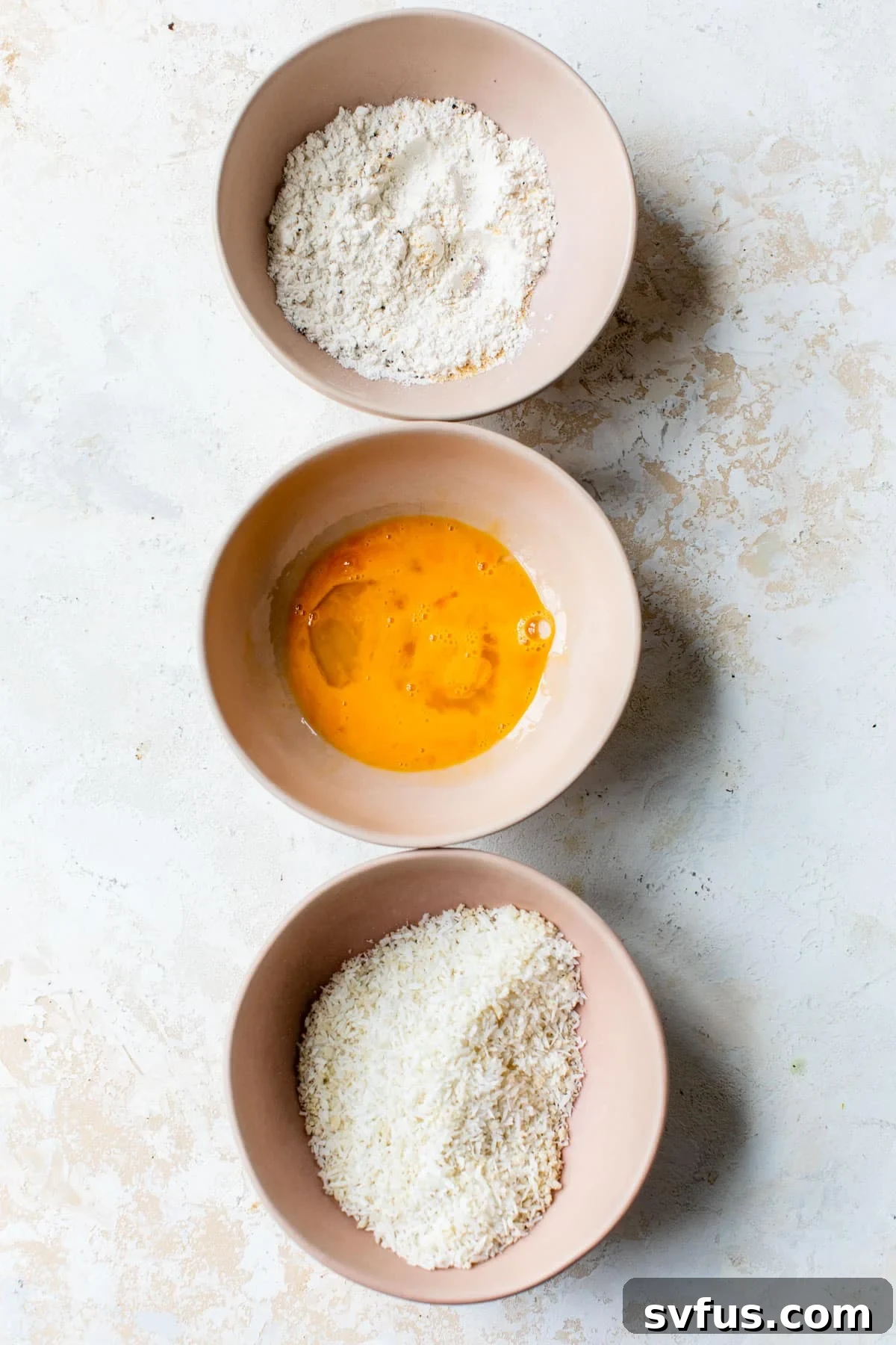 Three bowls in an assembly line: one with flour mixture, one with whisked eggs, and one with coconut flakes and panko breadcrumbs