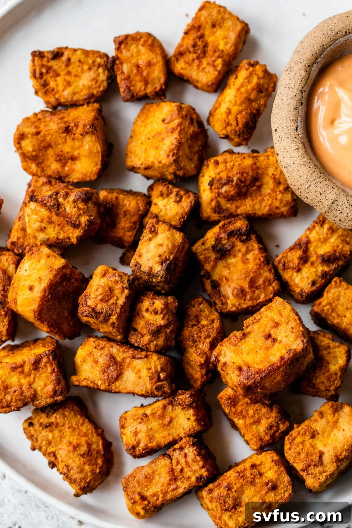 Close-up of golden, crispy air fryer tofu cubes on a white plate with a small bowl of dipping sauce beside it.
