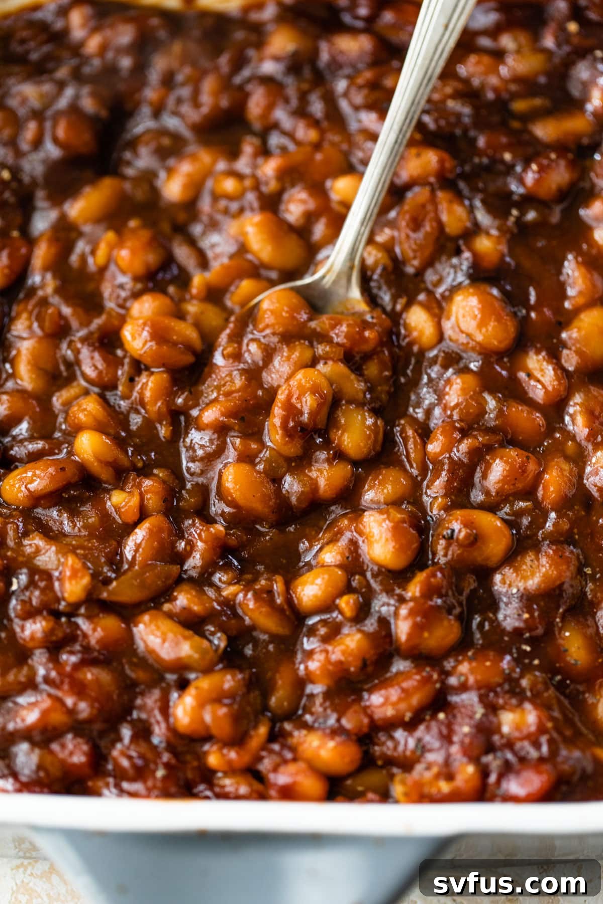 A baking dish filled with perfectly baked vegan beans, with a spoon nestled in, showing off their rich, thick sauce.