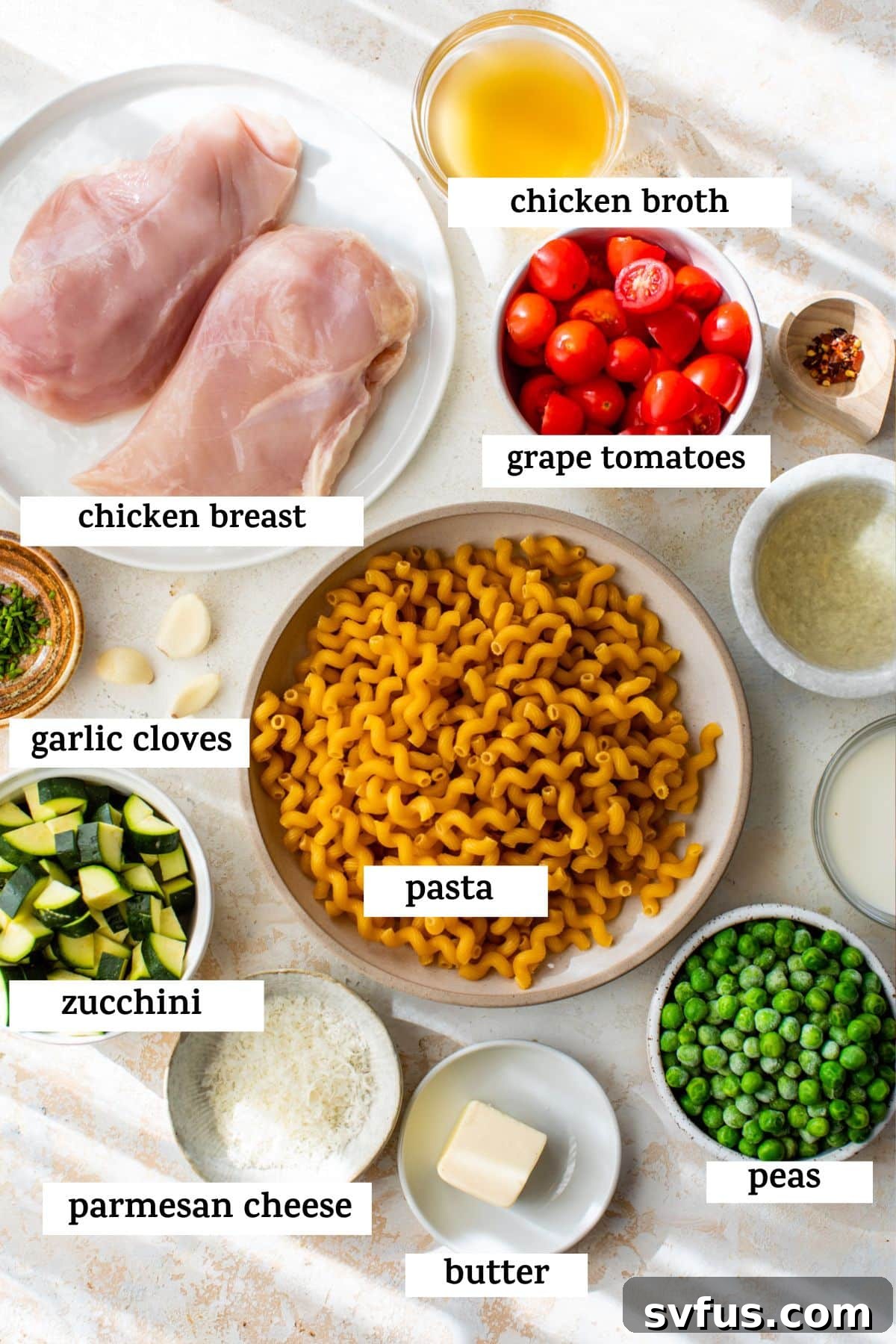 All fresh ingredients for chicken pasta primavera laid out on a wooden table, including chicken breast, zucchini, grape tomatoes, peas, garlic, pasta, chicken broth, white wine, half and half, and parmesan cheese.