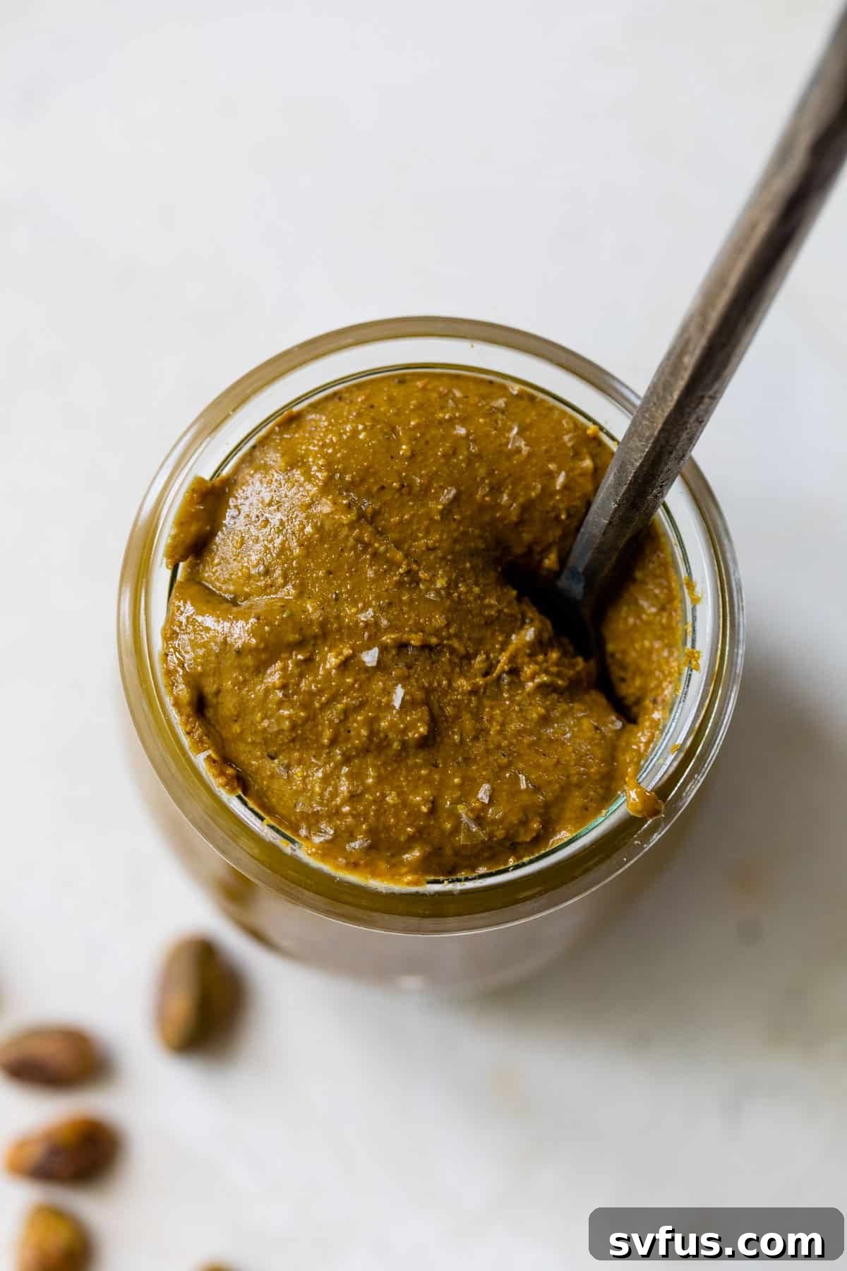 An overhead view of a glass jar full of homemade pistachio butter, with a small knife inside, sitting on a light surface.