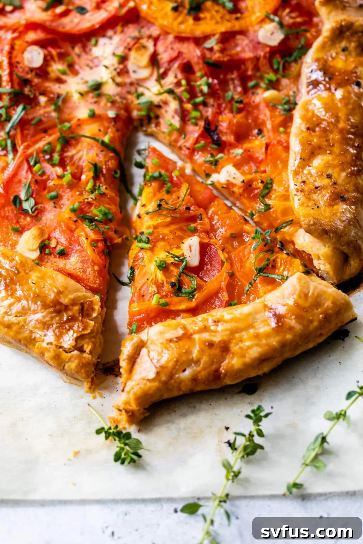 Close-up of a golden brown tomato galette garnished with fresh thyme and chives, with a slice removed, showcasing its delicious layers.