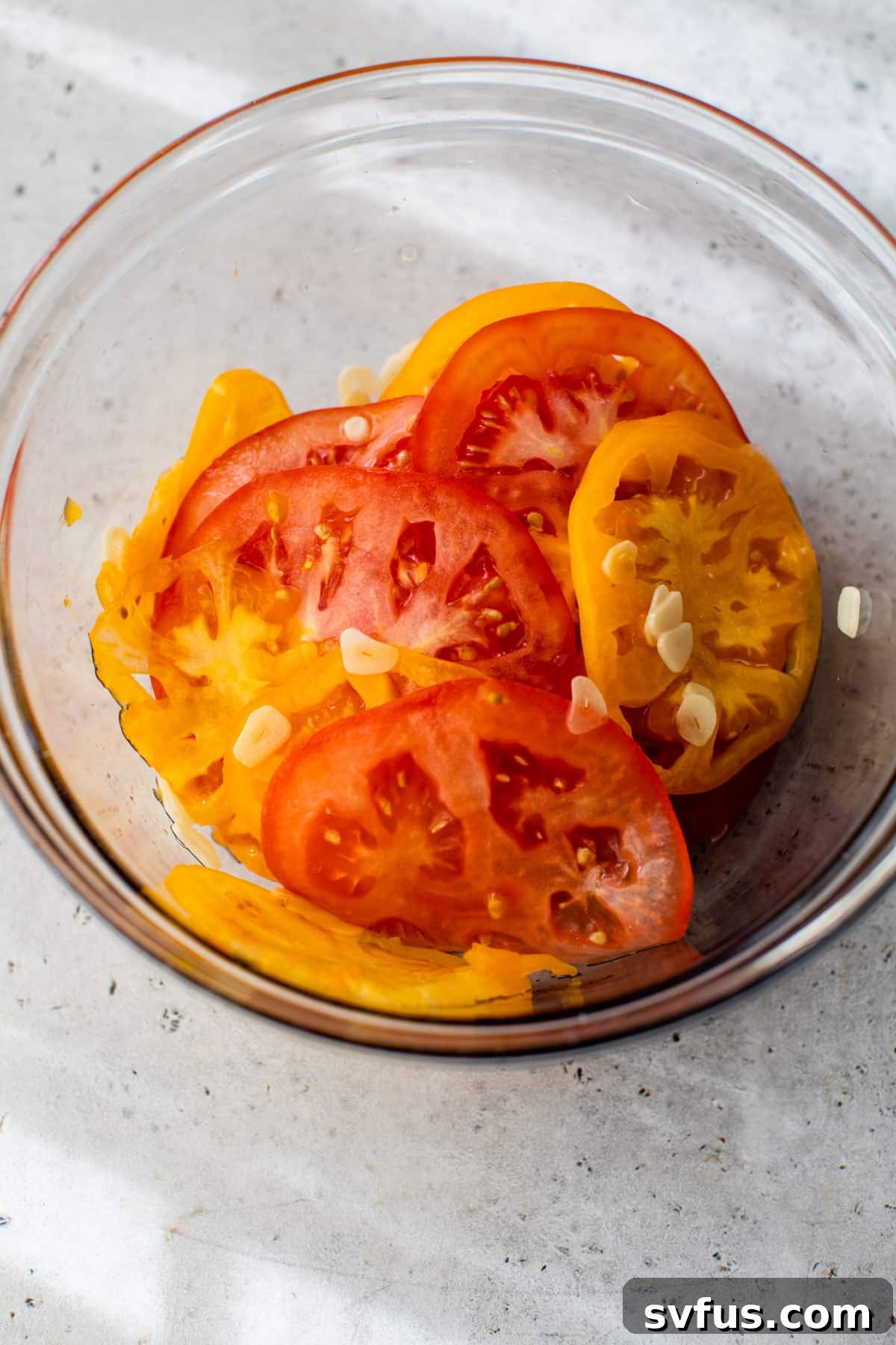 Sliced heirloom tomatoes mixed with thin garlic slices and salt in a clear glass bowl, preparing for draining.