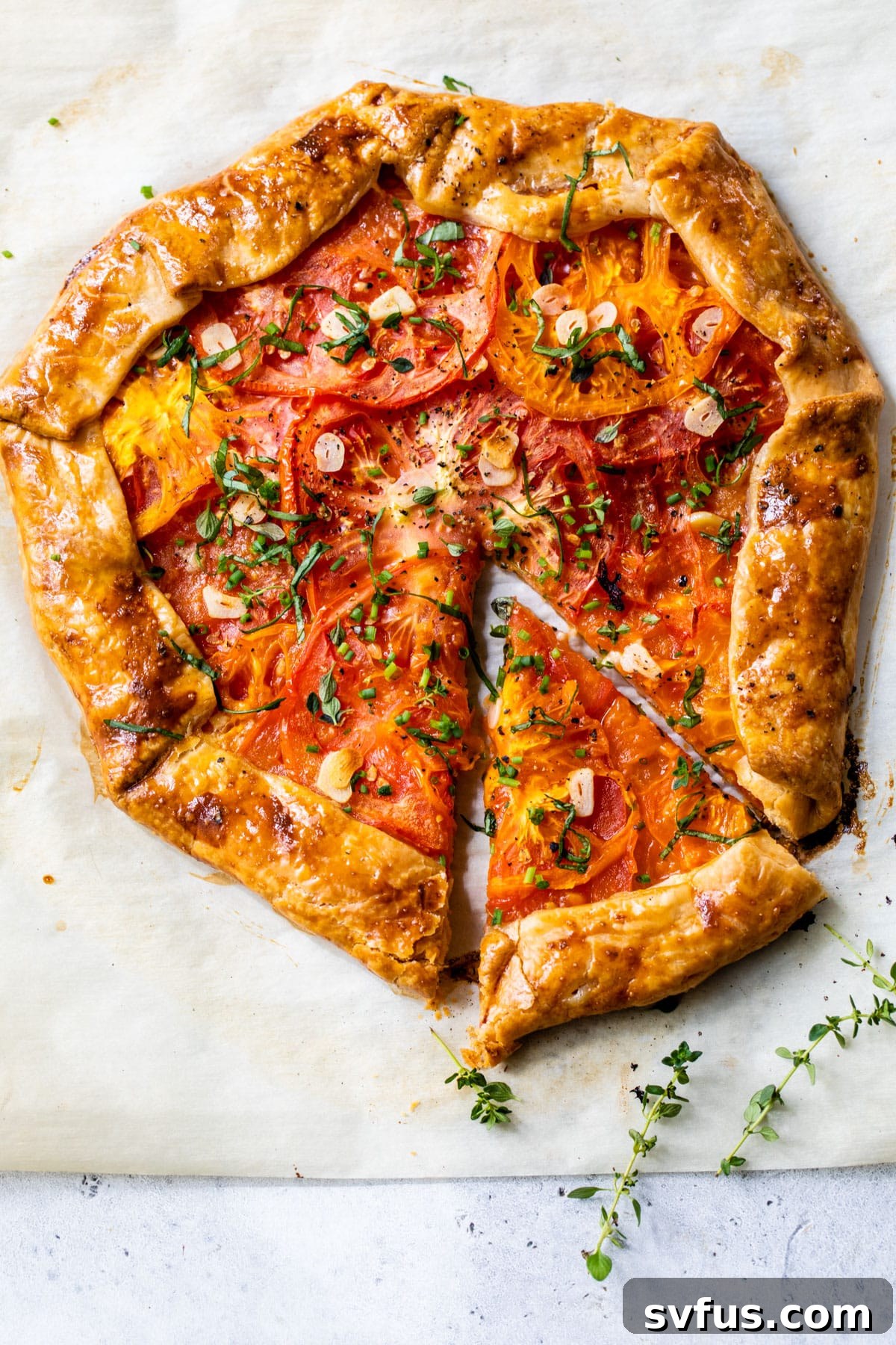 An overhead view of a baked tomato galette, garnished with fresh thyme and chives, with a slice already served.