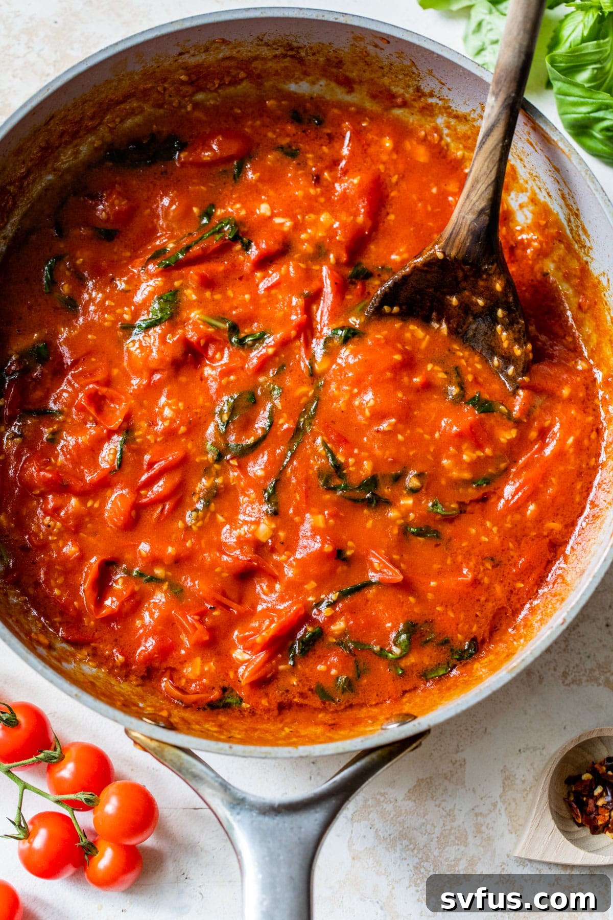 Close-up of vibrant cherry tomato pasta sauce simmering in a large skillet, with fresh basil, salt, and black pepper stirred in, creating an aromatic blend