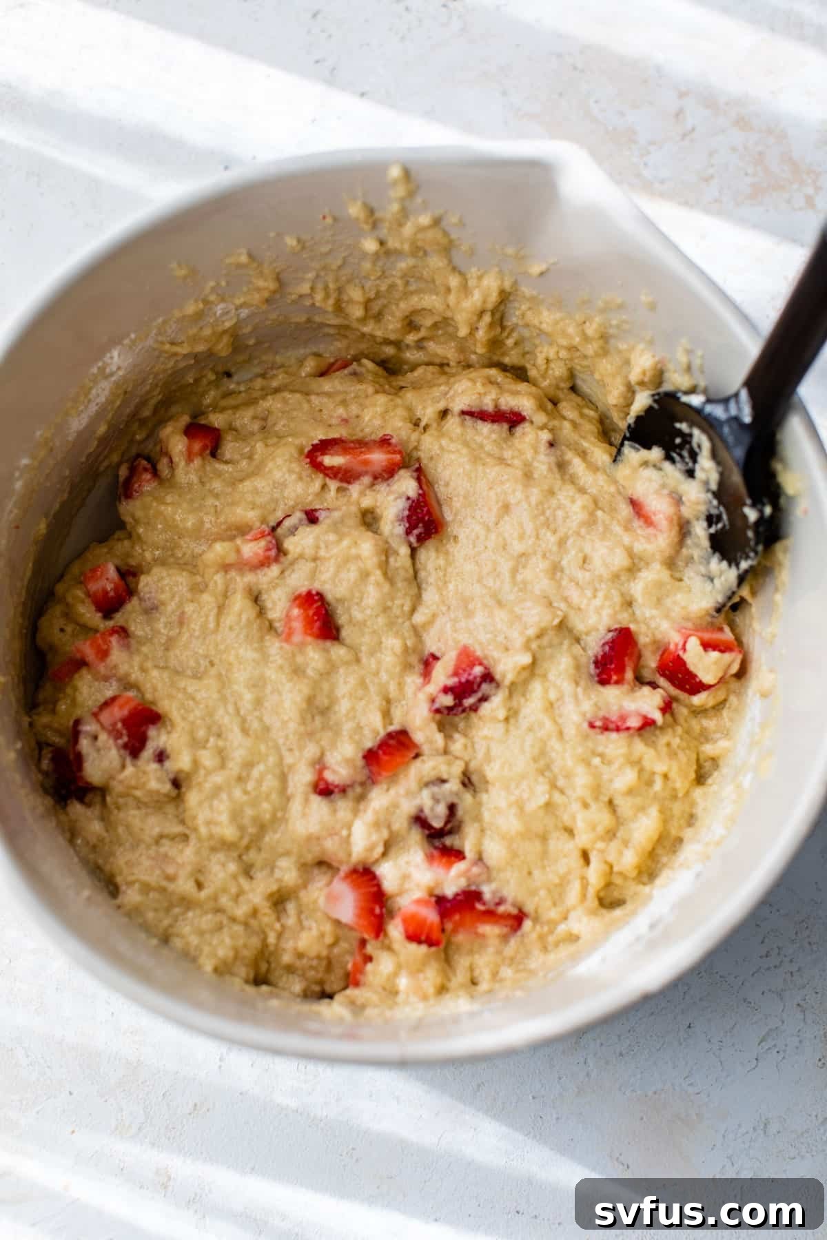Strawberry muffin batter in a large mixing bowl with a rubber spatula, showing the folded-in strawberry pieces.