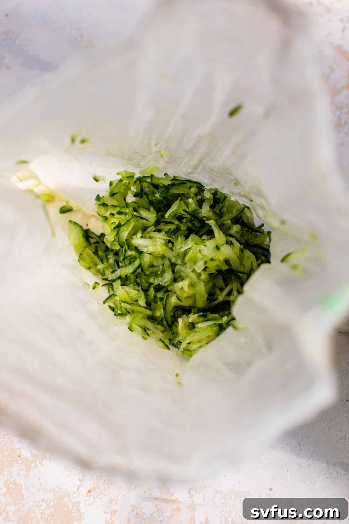 Close-up view of grated cucumber being squeezed between layers of paper towels to remove excess moisture, highlighting a crucial step in preparing tzatziki.