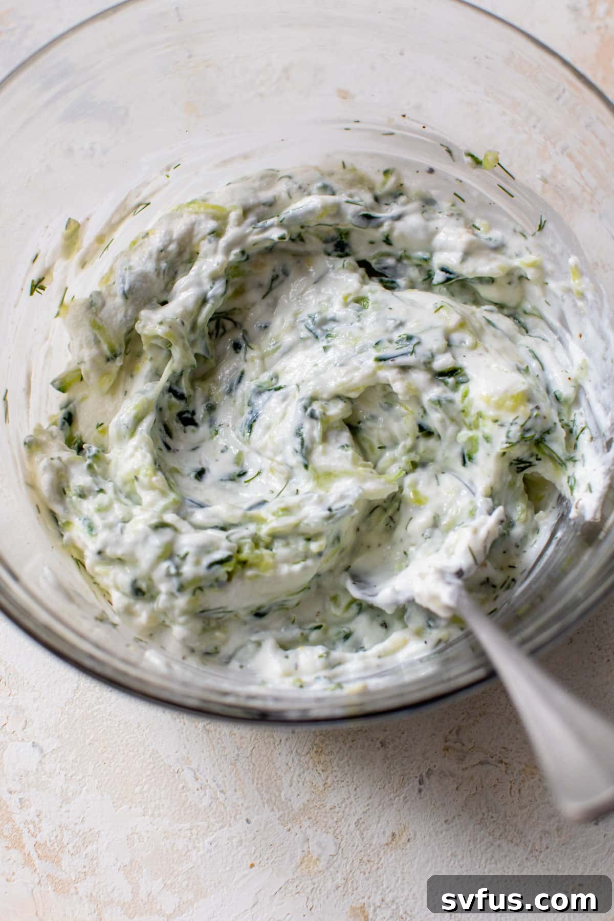 All tzatziki ingredients, including grated cucumber, Greek yogurt, garlic, dill, and lemon juice, being mixed together in a clear glass bowl with a spoon.