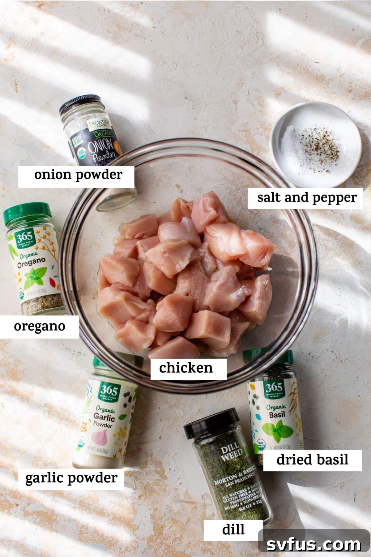 A collection of fresh ingredients for Greek chicken skewers laid out on a table, including raw chicken pieces, dried oregano, onion powder, garlic powder, dill, dried basil, salt, and pepper.
