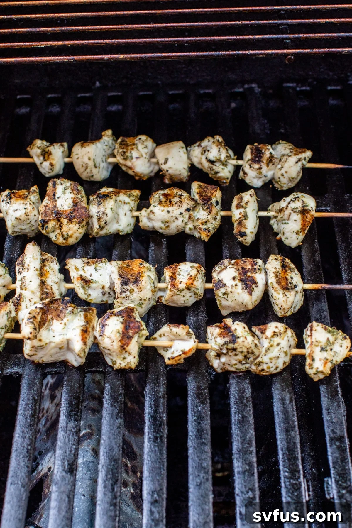 Several Greek chicken skewers sizzling on the hot grates of an outdoor grill, with visible char marks developing on the chicken pieces.