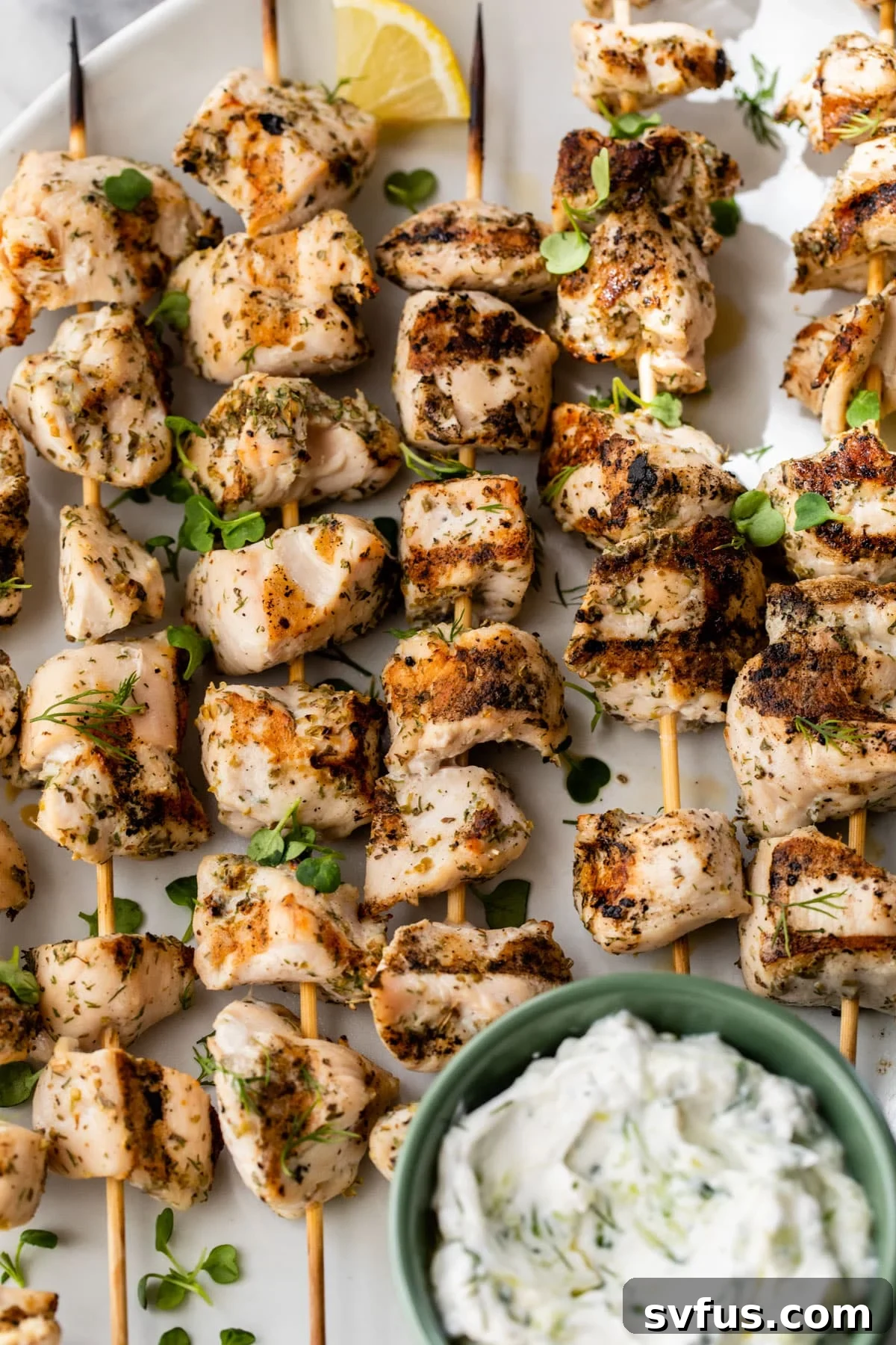 A close-up shot of perfectly grilled Greek chicken skewers on a plate, accompanied by a small bowl of creamy tzatziki sauce, garnished with fresh herbs.