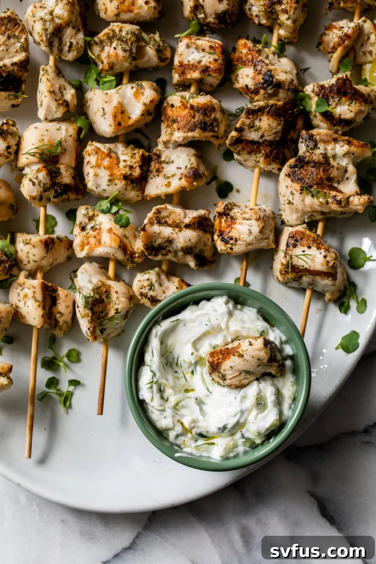 Close-up of a single Greek chicken skewer with a piece of chicken dipped into creamy tzatziki sauce on a white plate, showcasing the tenderness and flavor.