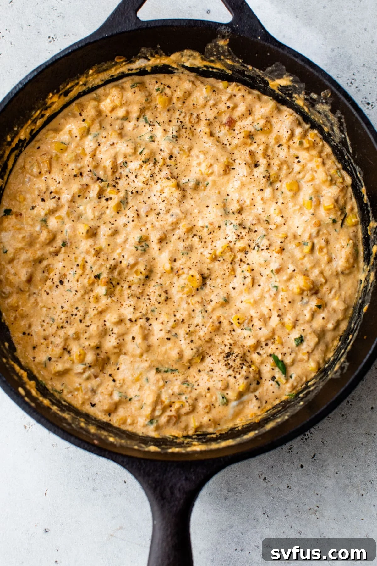 Cream cheese and Greek yogurt being stirred into the corn mixture in a cast iron skillet, creating a smooth dip.