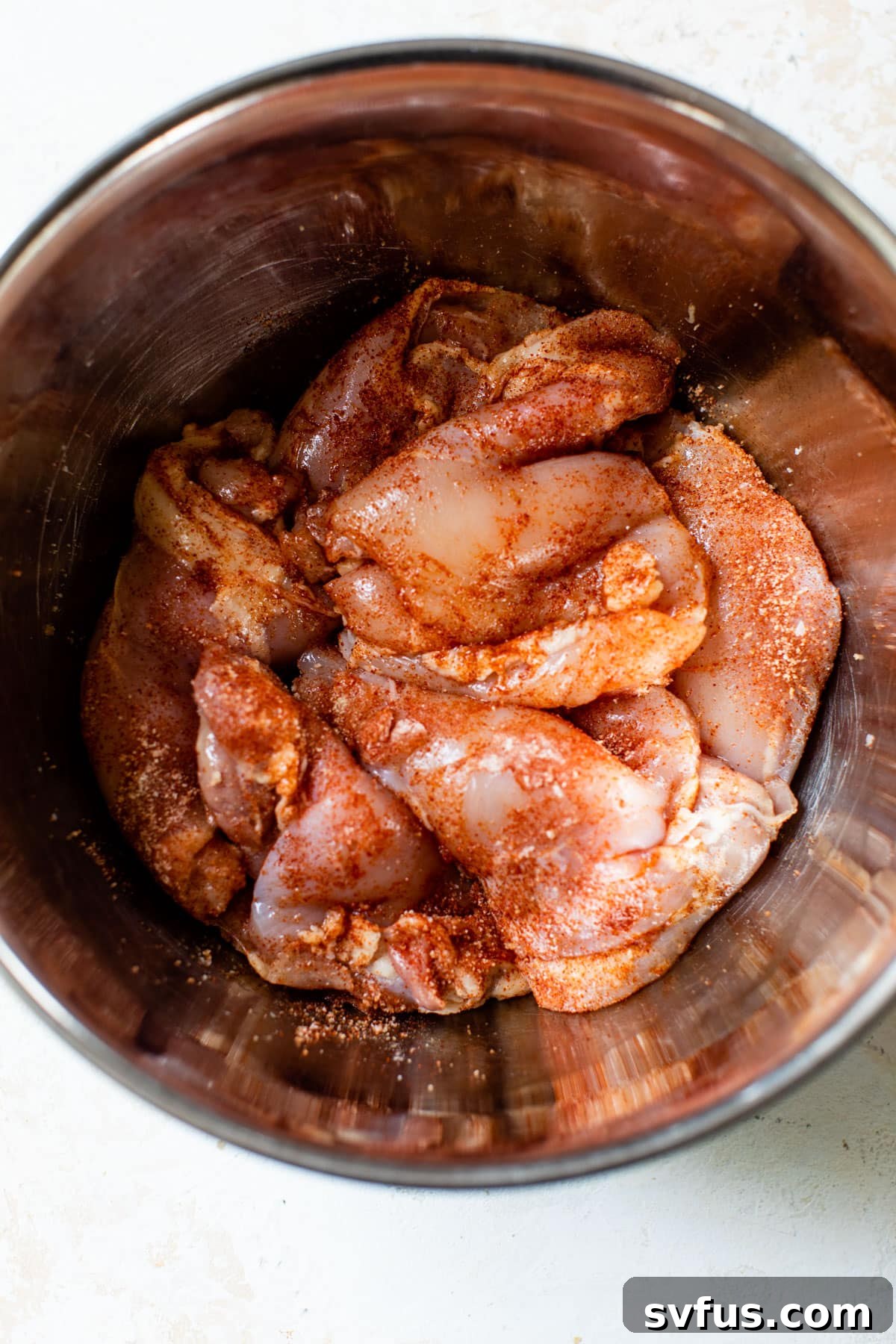 raw chicken thighs in a metal bowl with seasonings on top, ready for mixing