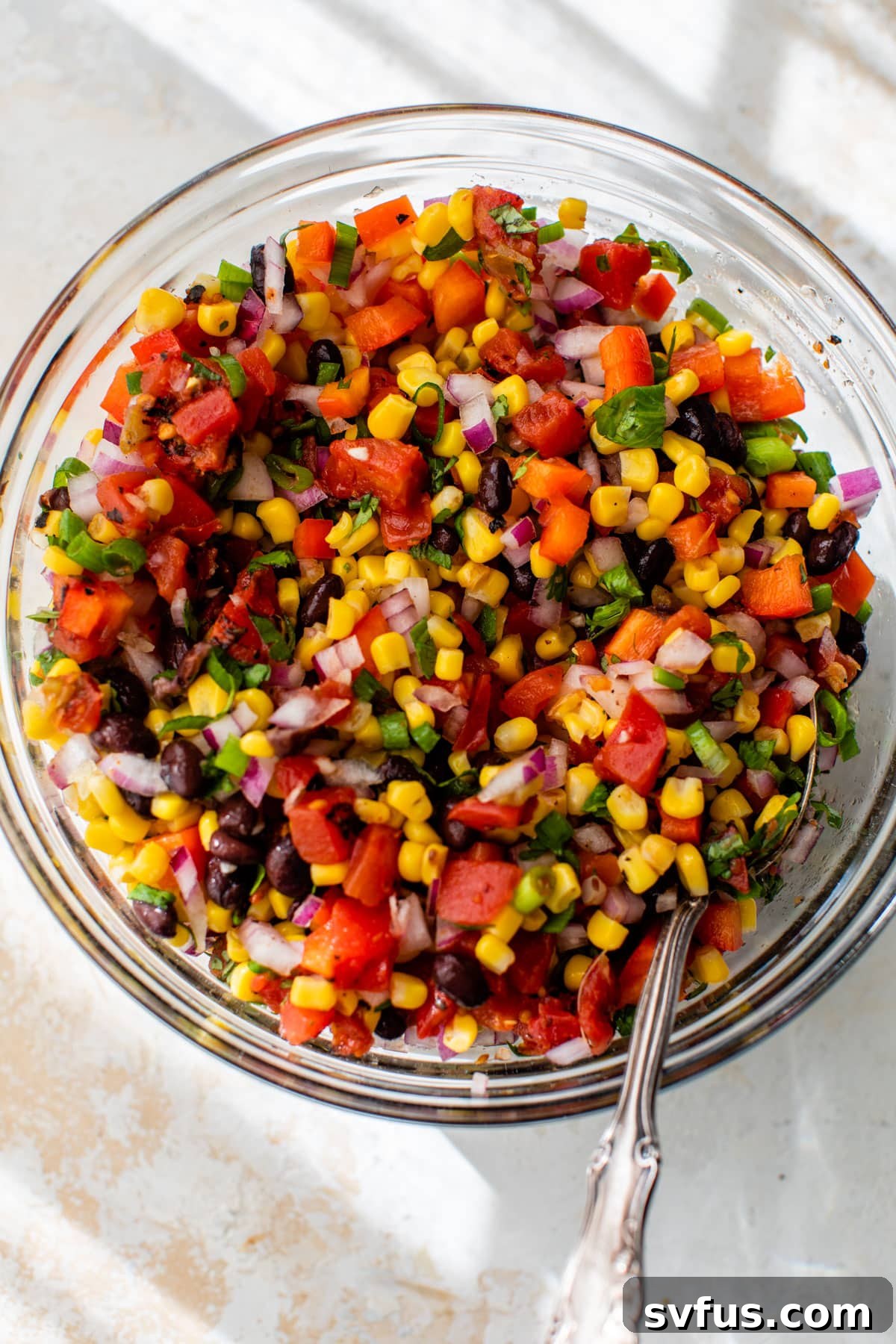 All the diced and prepared ingredients for the salsa, including black beans, corn, tomatoes, onions, bell pepper, and cilantro, are tossed together in a large clear glass bowl with a serving spoon.
