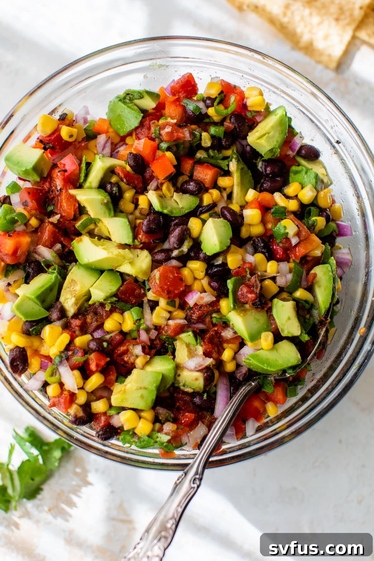 Diced avocado has been added to the glass bowl containing the mixed salsa ingredients and dressing, showing the final stage of preparation before serving.