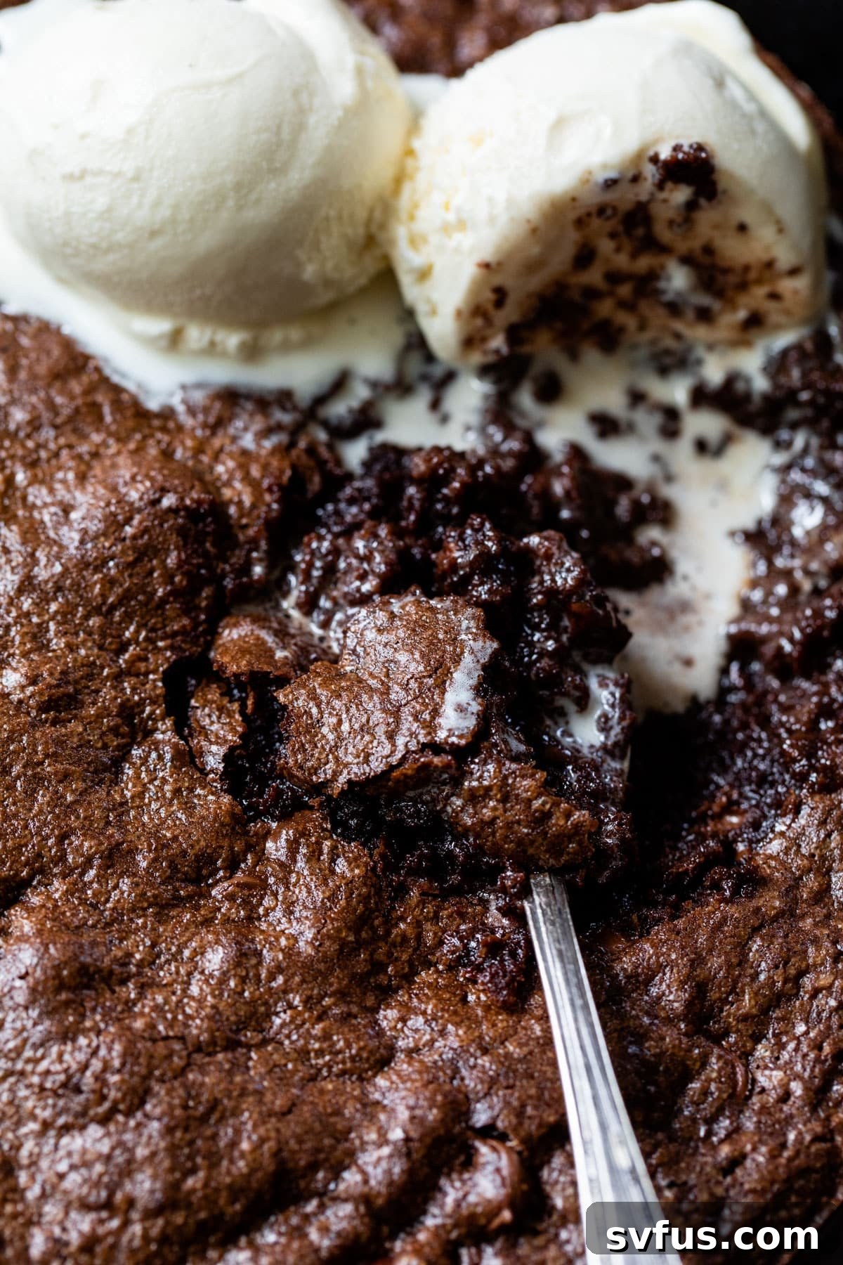 Close-up of a warm brownie skillet with two scoops of melting vanilla ice cream and a spoon, inviting a delicious bite.