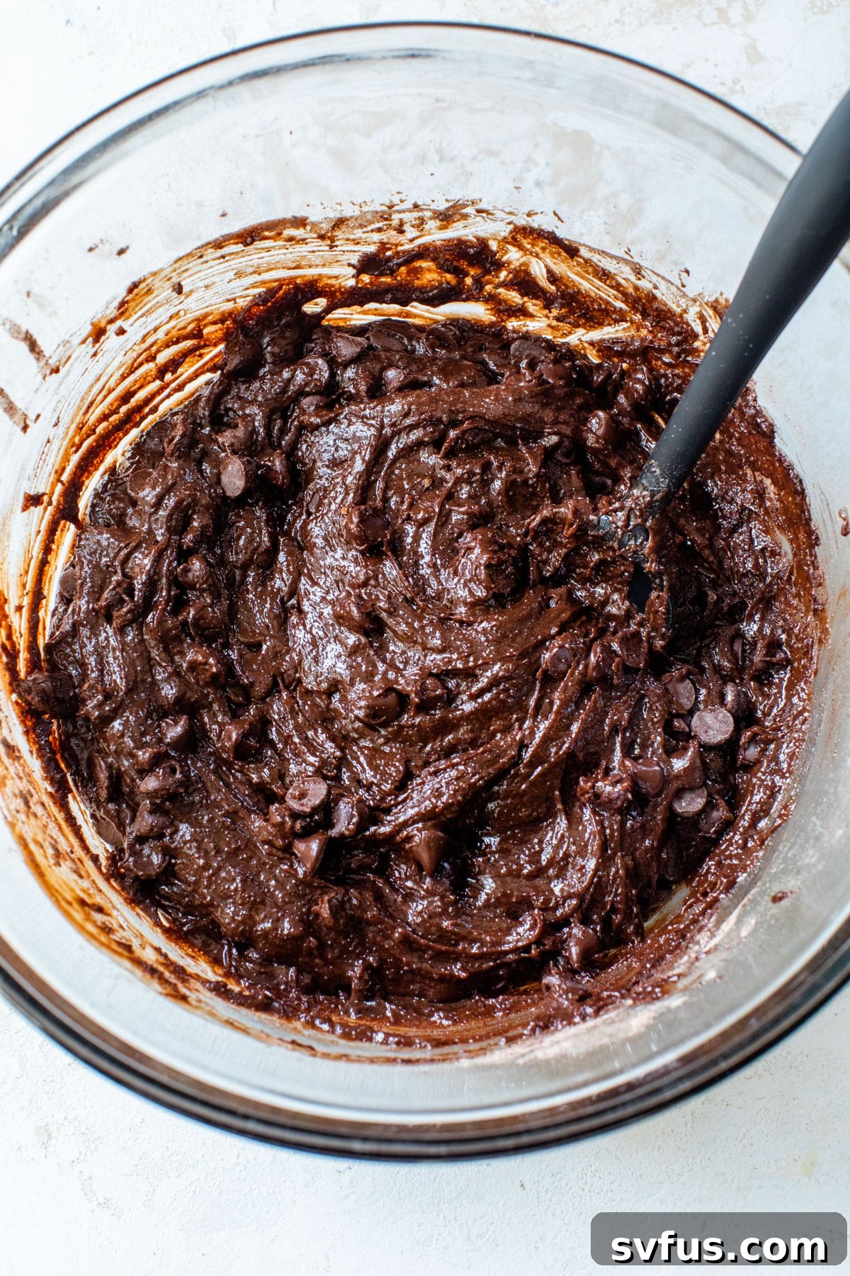 Thick brownie skillet batter with visible chocolate chips in a glass mixing bowl, alongside a spatula.