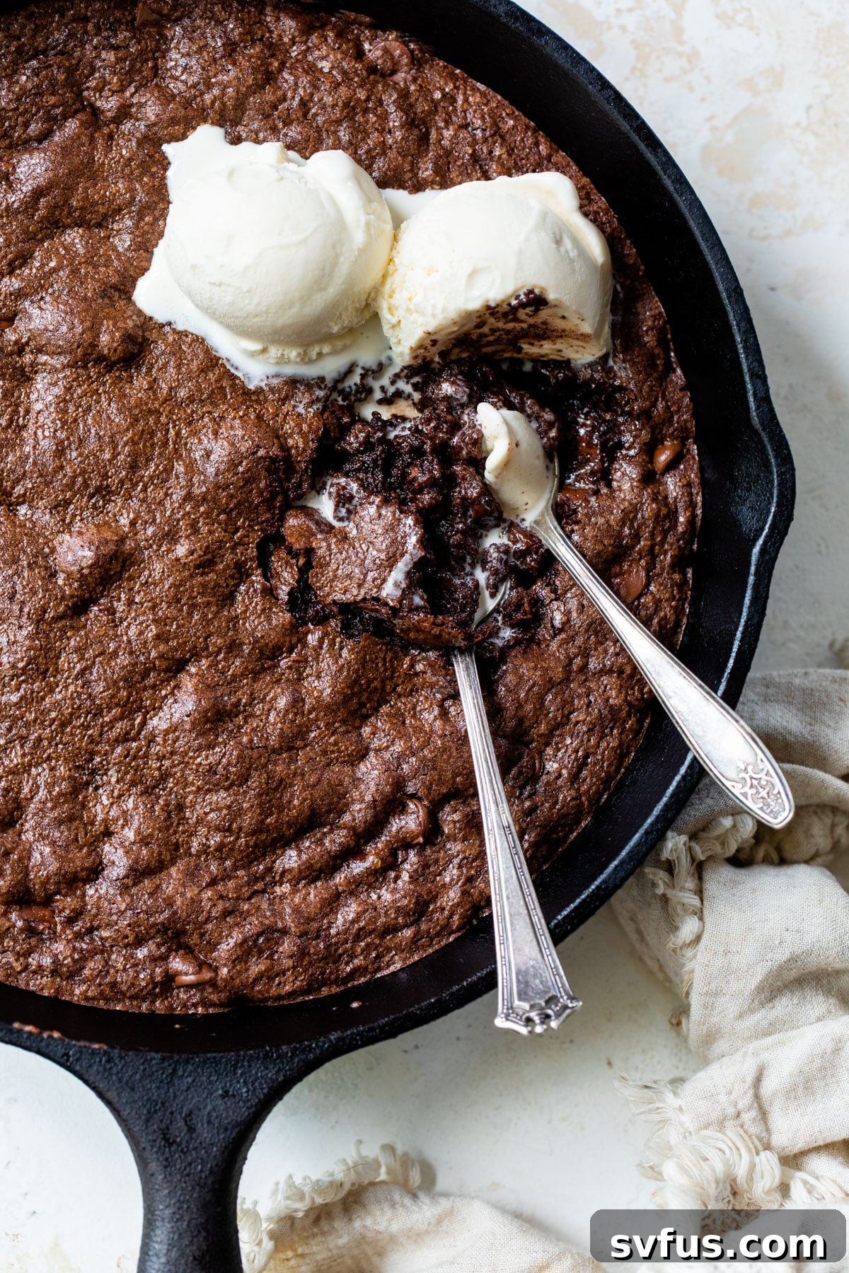 An inviting overhead shot of a warm brownie skillet adorned with two scoops of vanilla ice cream and two spoons, ready for sharing.