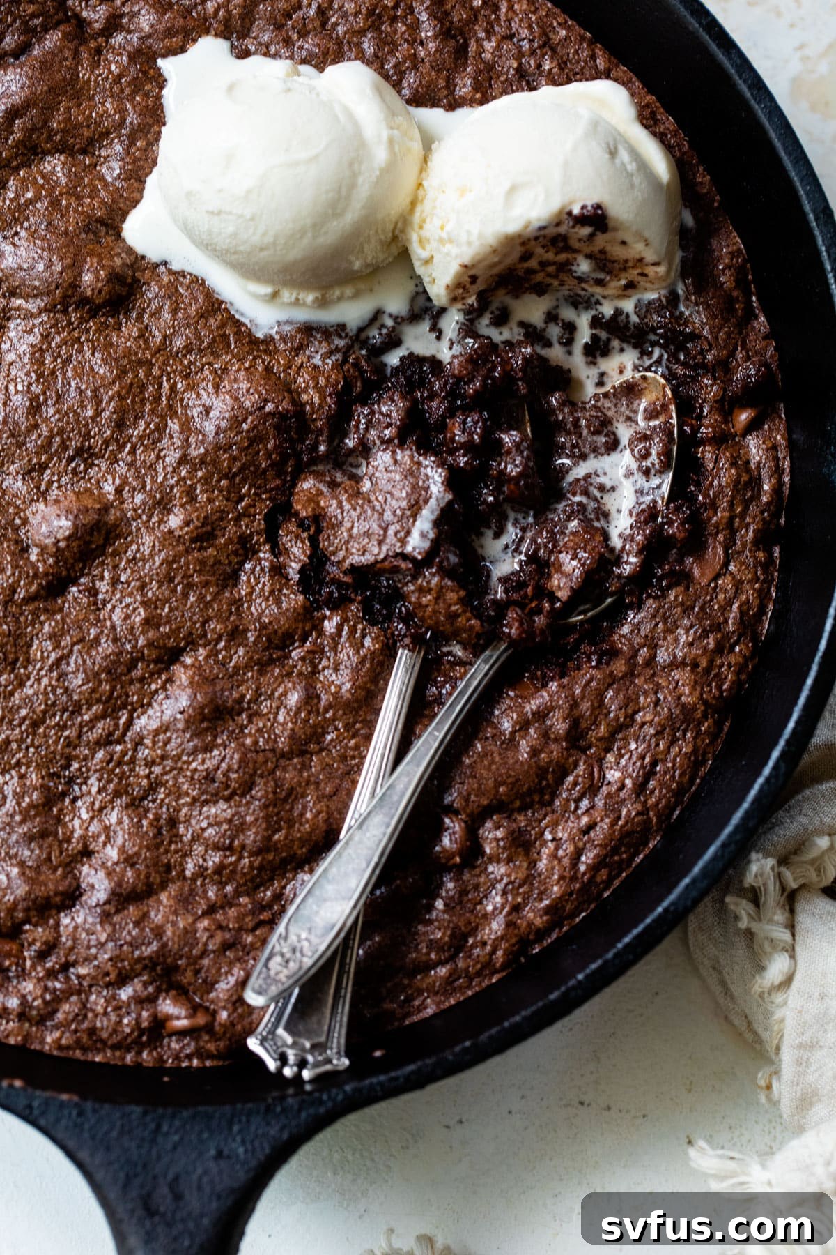An inviting overview of a rich brownie skillet, perfectly baked, adorned with two scoops of creamy vanilla ice cream and two spoons, ready for indulgence.