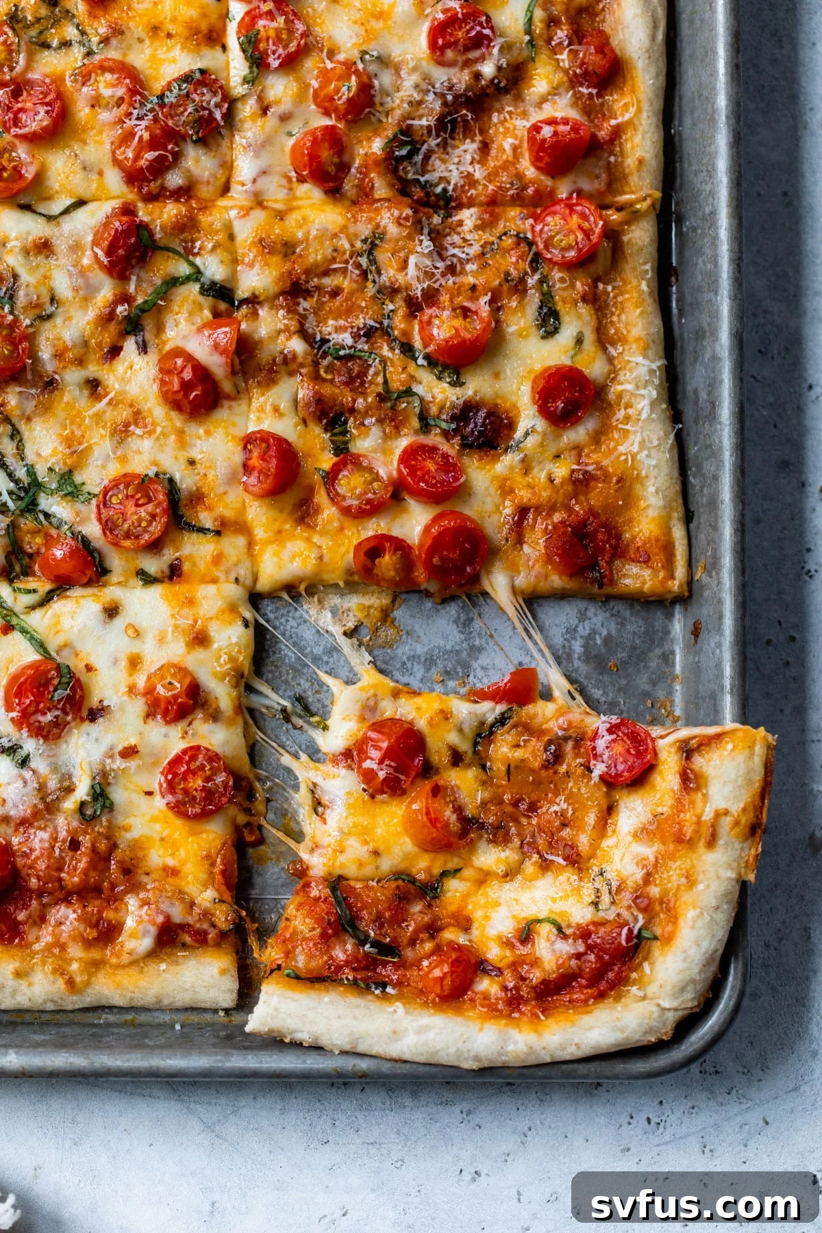 A perfectly baked sheet pan pizza with a slice being lifted, showing melted cheese and fresh basil.