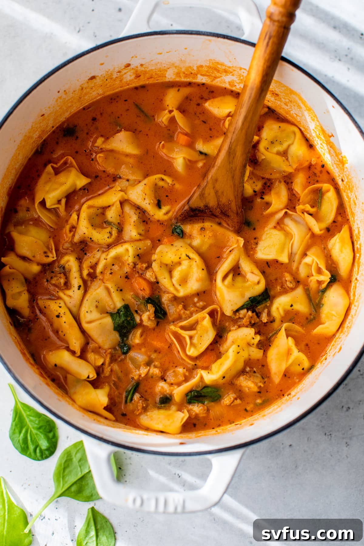 Sausage tortellini soup simmering in a Dutch oven with a wooden spoon resting inside.