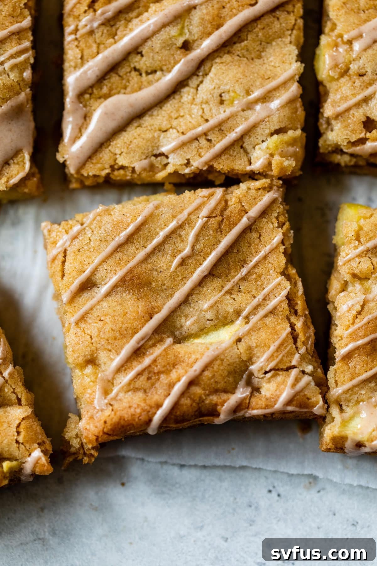 Close-up photograph of multiple sliced apple blondies on a surface, clearly showing the tender apple pieces embedded within the golden blondie and the sweet cinnamon glaze drizzled over the top.