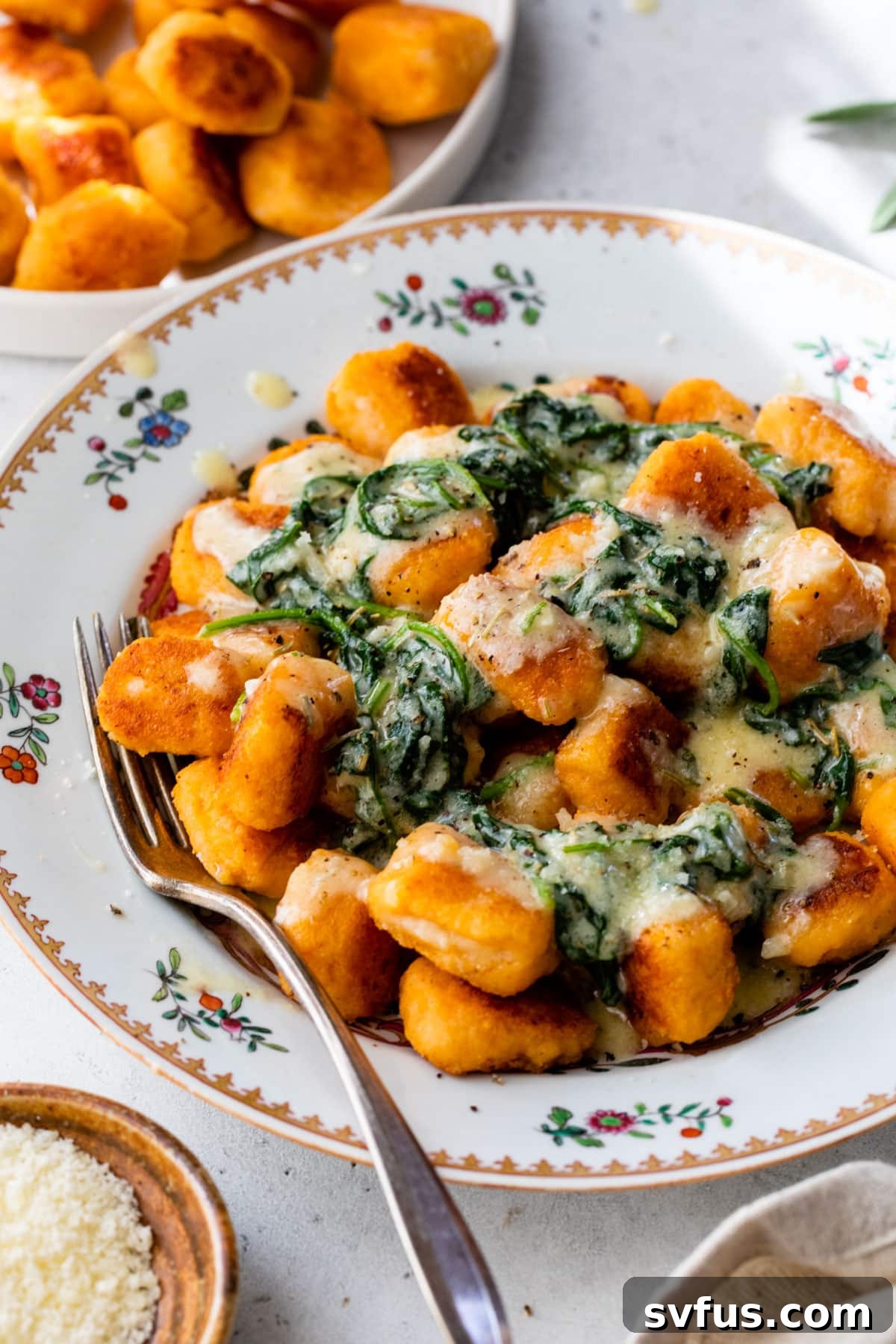 A beautifully presented bowl of sweet potato gnocchi, garnished with fresh cooked spinach and a dollop of rich butter sauce, with a fork gently piercing a piece.