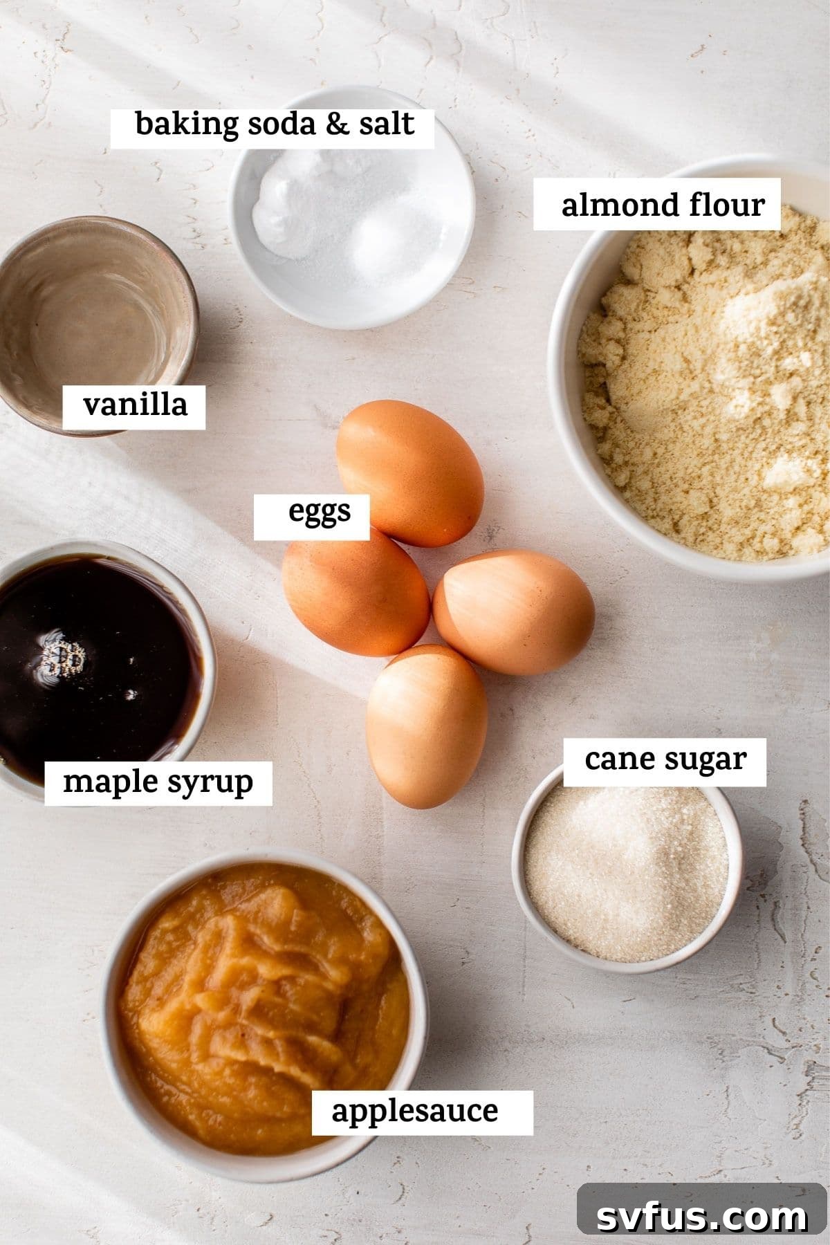 Various baking ingredients including almond flour, sugar, eggs, applesauce, and maple syrup laid out on a clean kitchen counter.