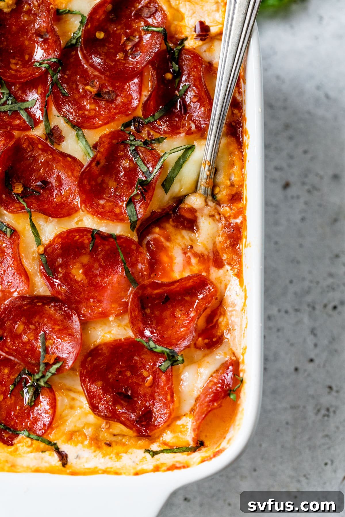 Close-up view of hot pizza dip in a baking dish with a serving spoon, topped with chopped fresh basil for garnish.