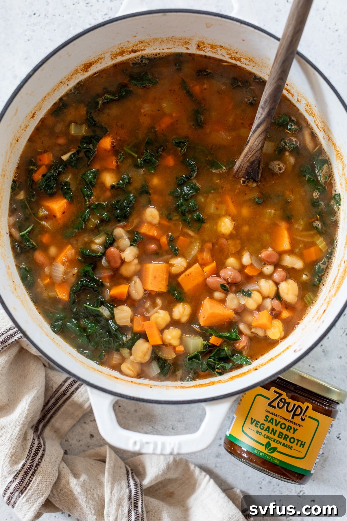 A Dutch oven full of simmering three bean soup, with a wooden spoon resting inside and a jar of vegan broth beside it.