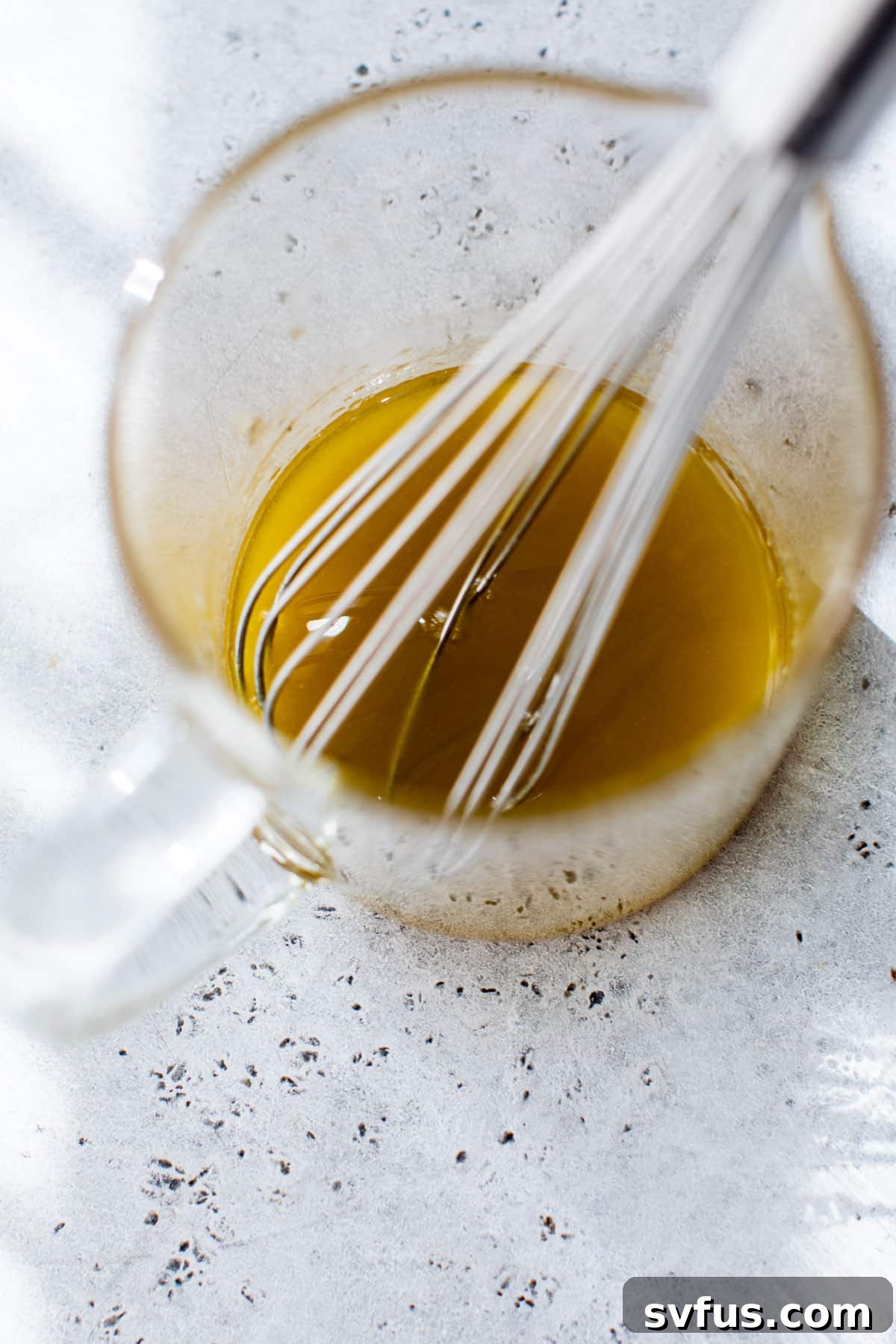 A glass measuring cup holds a beautifully whisked dressing, showcasing the blend of olive oil, apple cider vinegar, honey, and Dijon mustard.