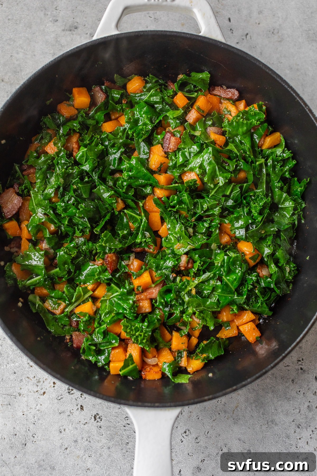 Chopped kale being added to a mixture of diced sweet potatoes and bacon in a cast iron skillet, ready for sautéing.