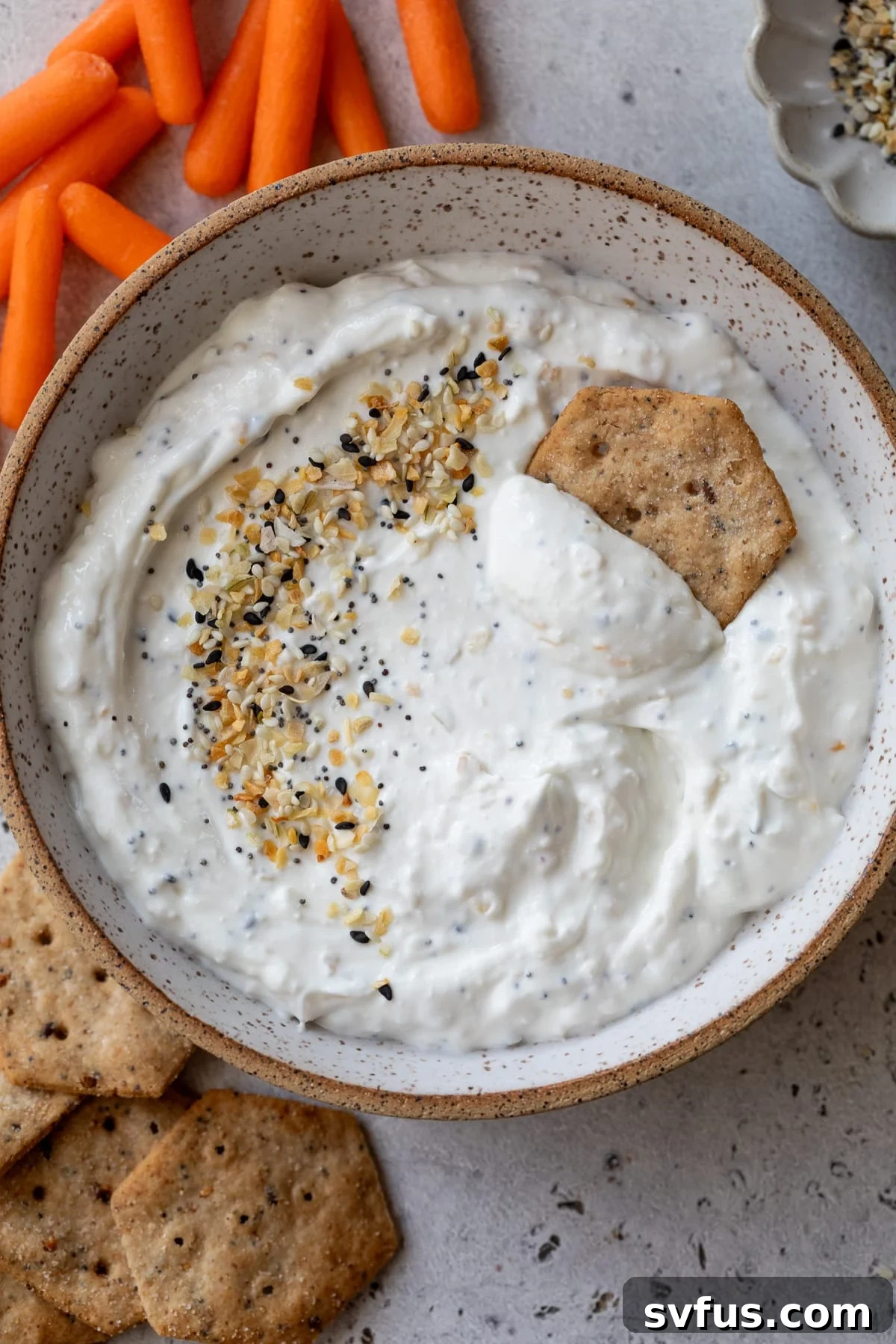 everything bagel dip in a bowl with a cracker and carrots off to the side, showcasing its creamy texture and vibrant seasoning.