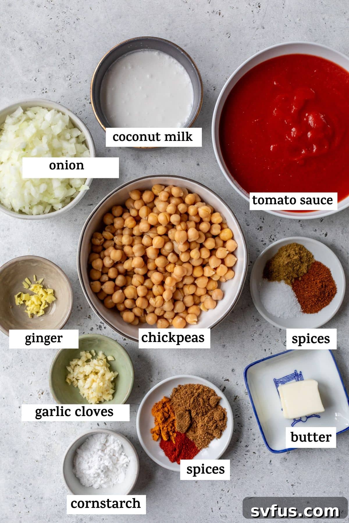 A flat lay of fresh ingredients laid out on a table: a diced onion, a can of full-fat coconut milk, tomato sauce, fresh ginger, canned chickpeas, an assortment of Indian spices, garlic cloves, vegan butter, and cornstarch, all essential for butter chickpeas.