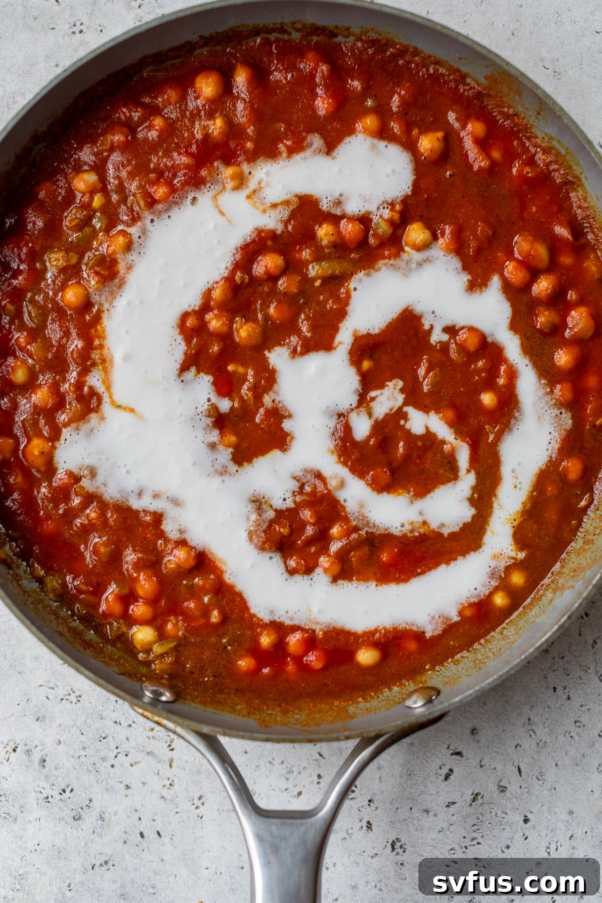 A close-up shot of a large skillet filled with simmering butter chickpeas and tomato sauce, as creamy coconut milk is being poured in, ready to be stirred and thicken the curry.