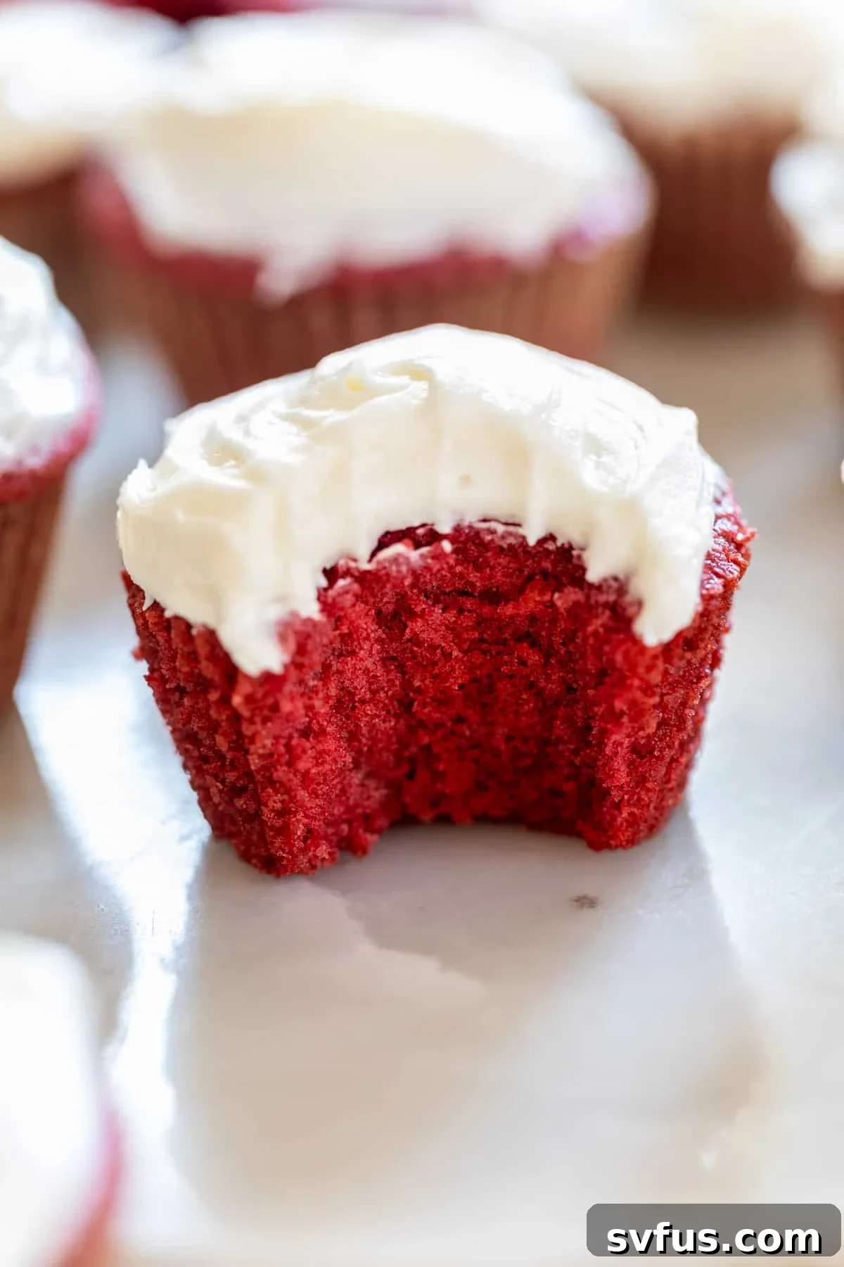 A perfectly baked, vibrant red velvet cupcake with a swirl of cream cheese frosting, with a bite taken out revealing its moist interior.