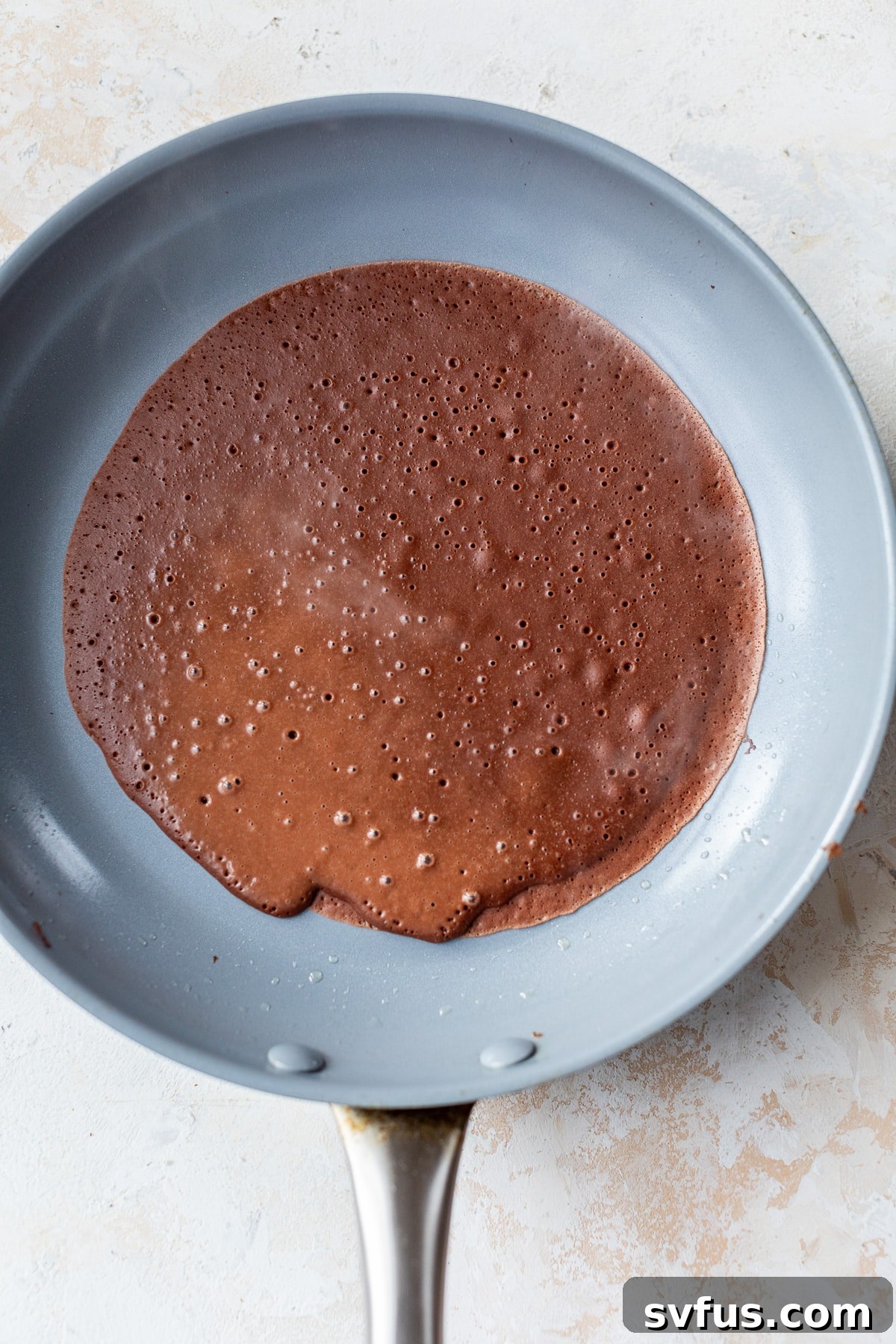 chocolate crepe batter being added to a skillet and swirled to cover the surface