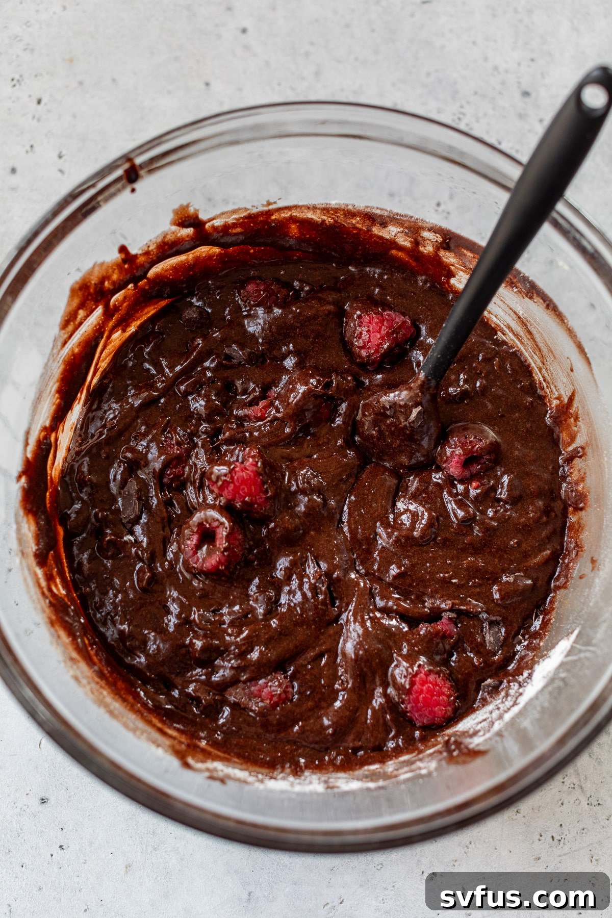 Brownie batter with fresh raspberries folded in, sitting in a glass bowl with a spatula.