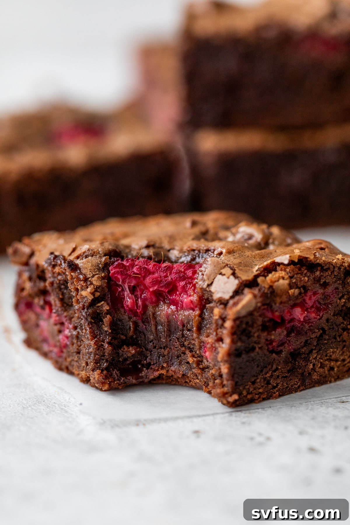 Close-up side view of a chocolate raspberry brownie with a bite taken out, showing its fudgy interior and melted chocolate pools.