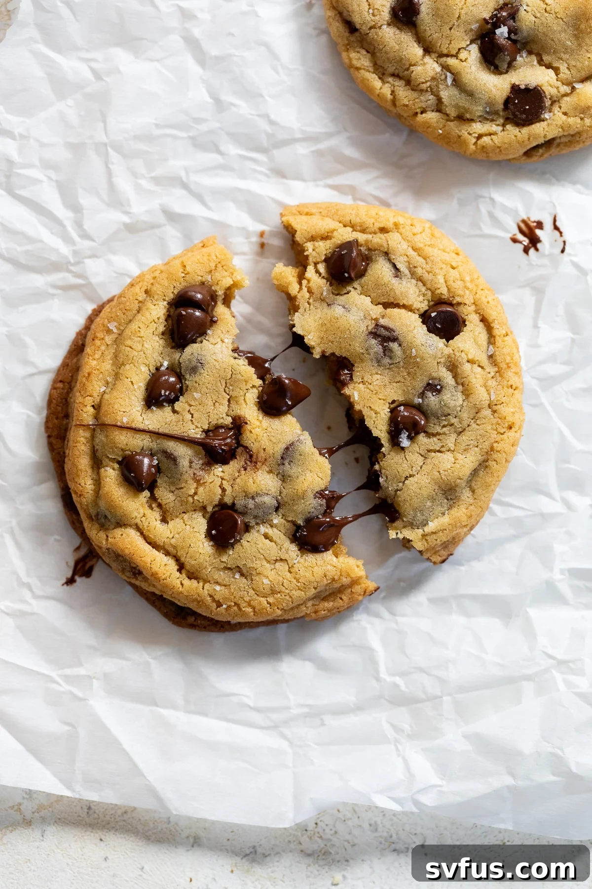 A single-serve chocolate chip cookie split in half, showcasing its gooey center, resting on parchment paper.