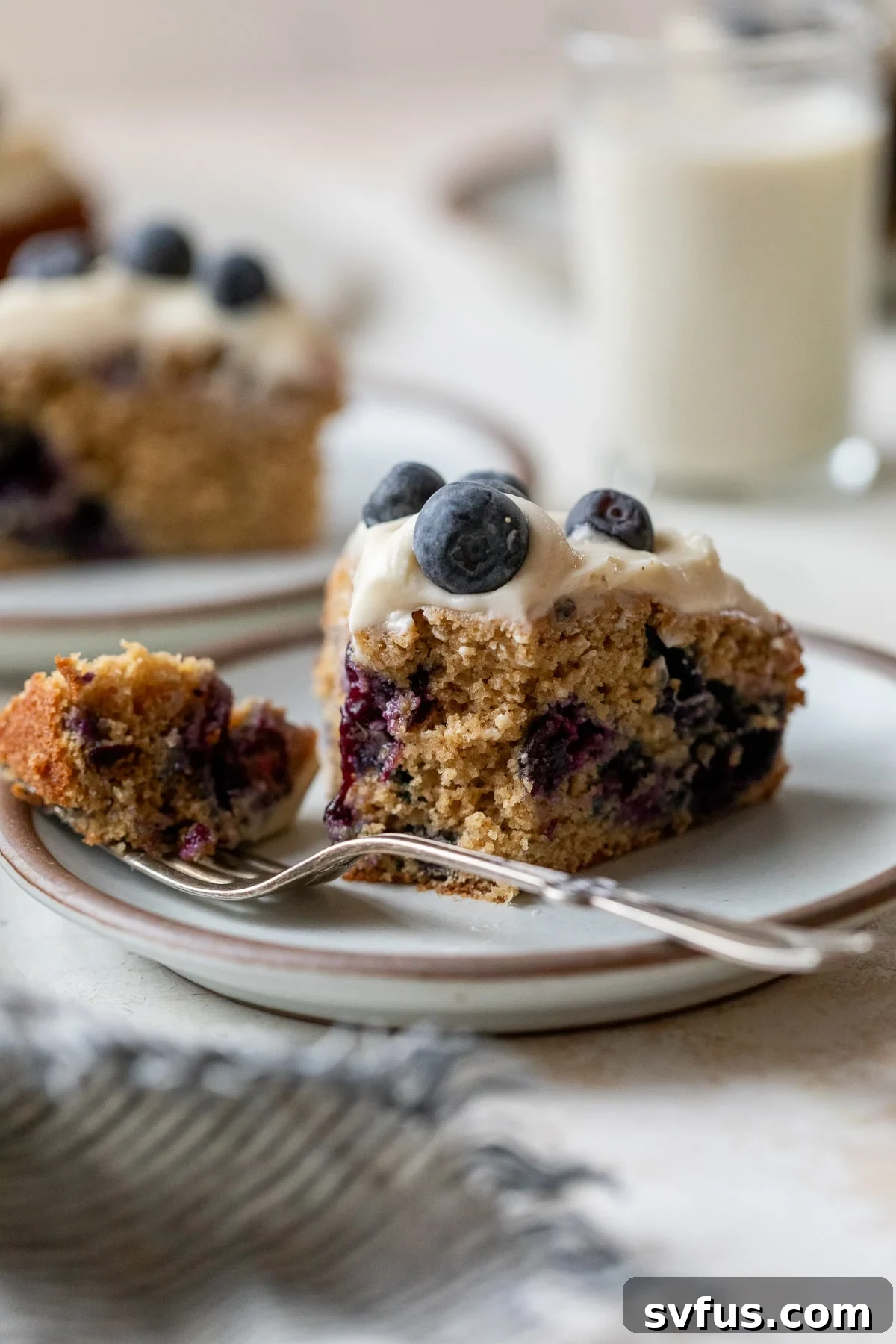 side view of a slice of blueberry cake on a plate with a fork and with a bite taken out