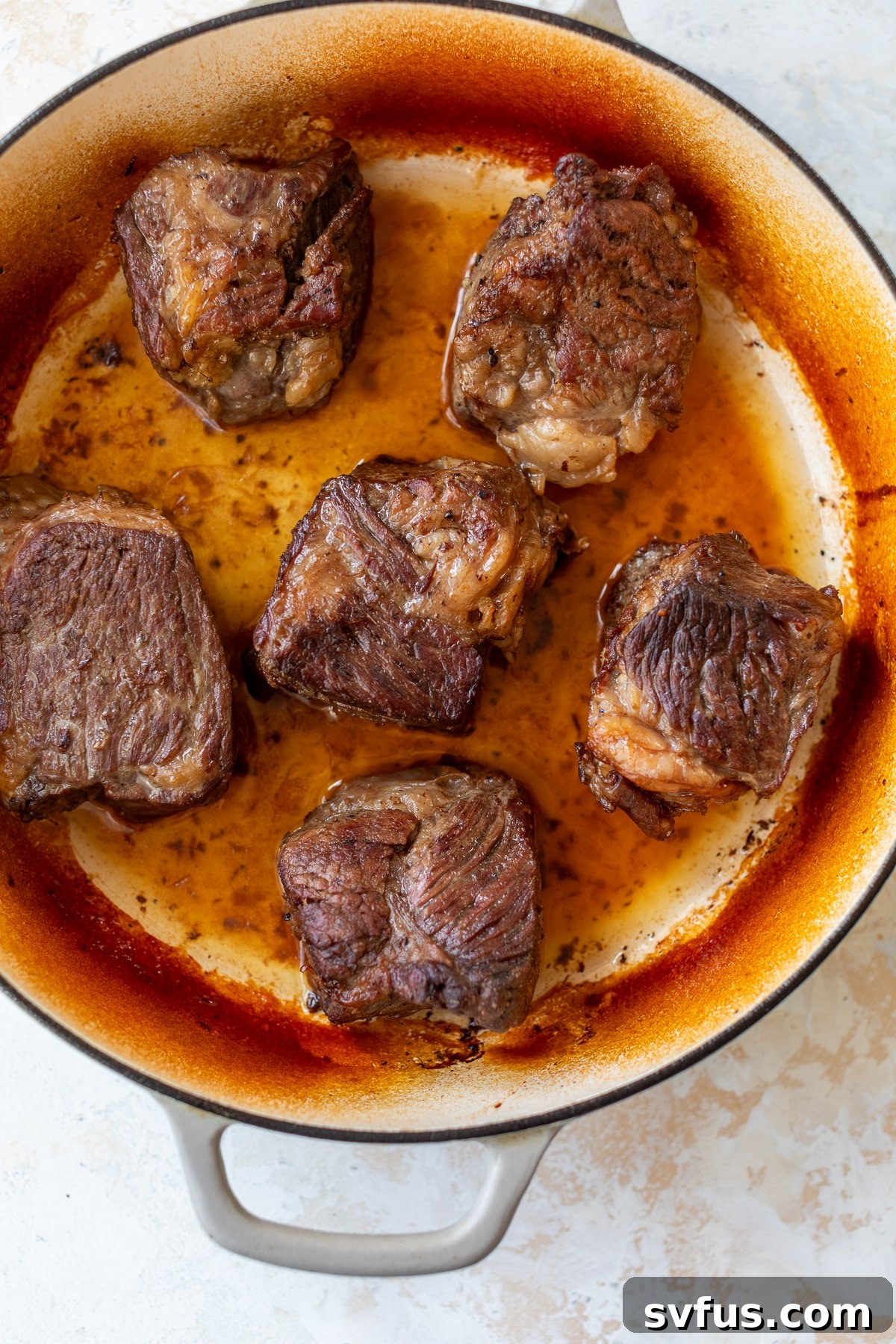 Seared boneless short ribs nestled back into a Dutch oven with beef broth, covered with a lid, ready for oven braising.