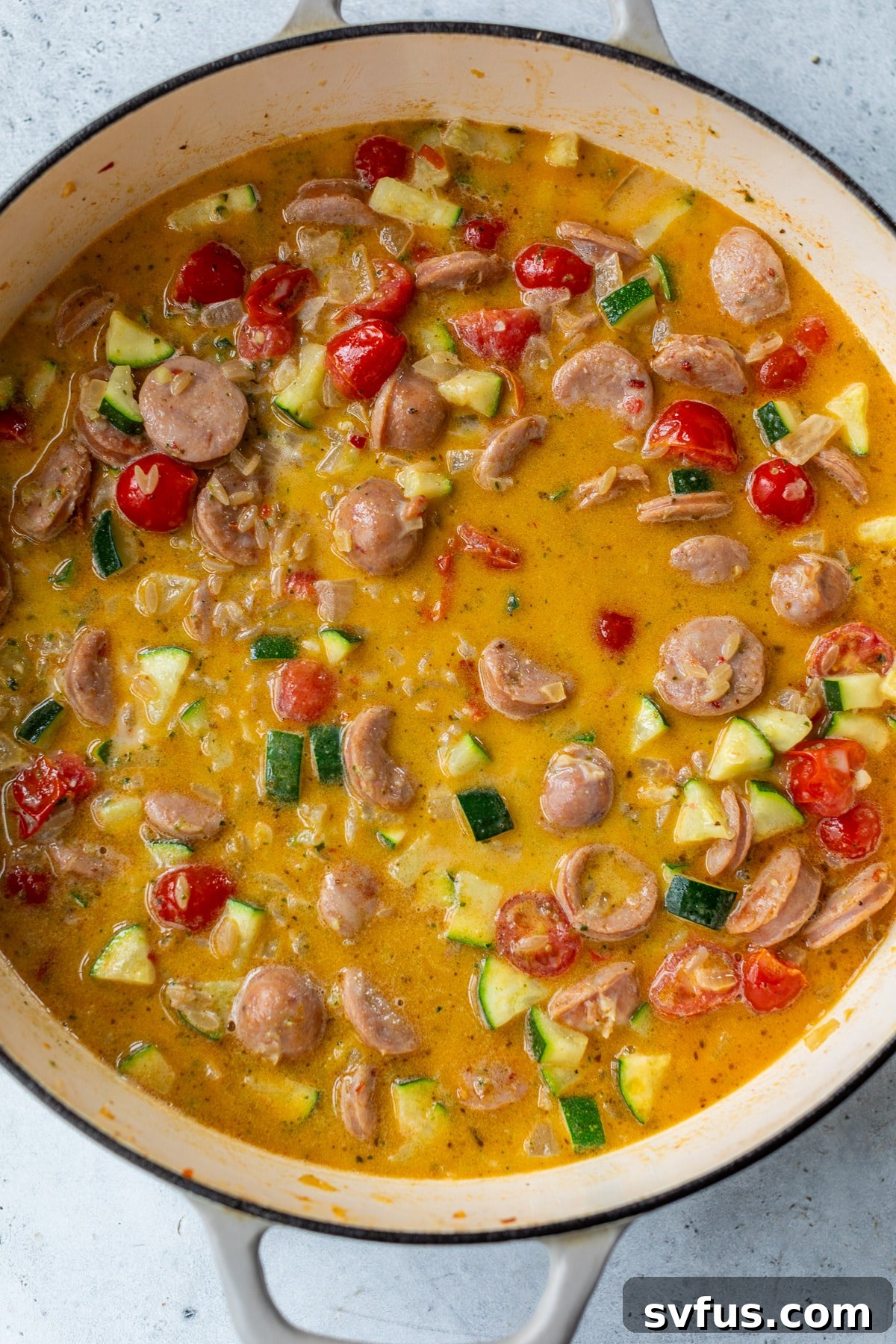 Chicken broth and heavy cream being poured into the Dutch oven with chicken sausage and vegetables, ready for orzo to cook.