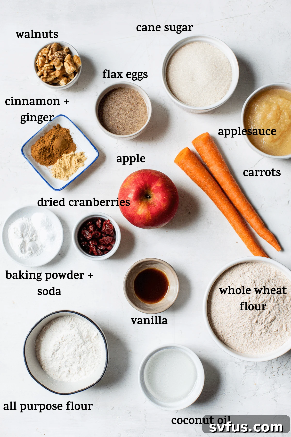 Various muffin ingredients, including whole wheat flour, cane sugar, spices, carrots, apples, walnuts, and dried cranberries, neatly arranged on a white table.
