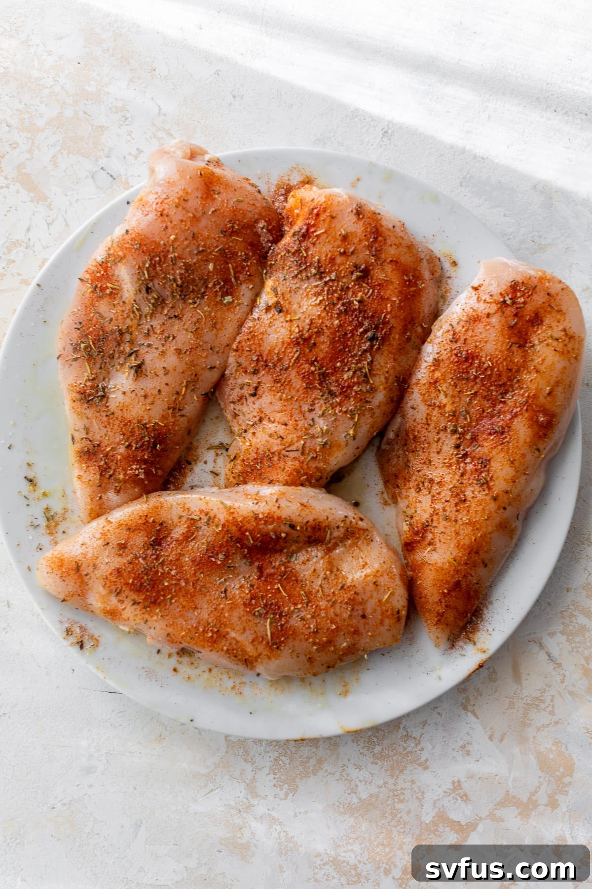 4 raw seasoned chicken breasts on a white plate.