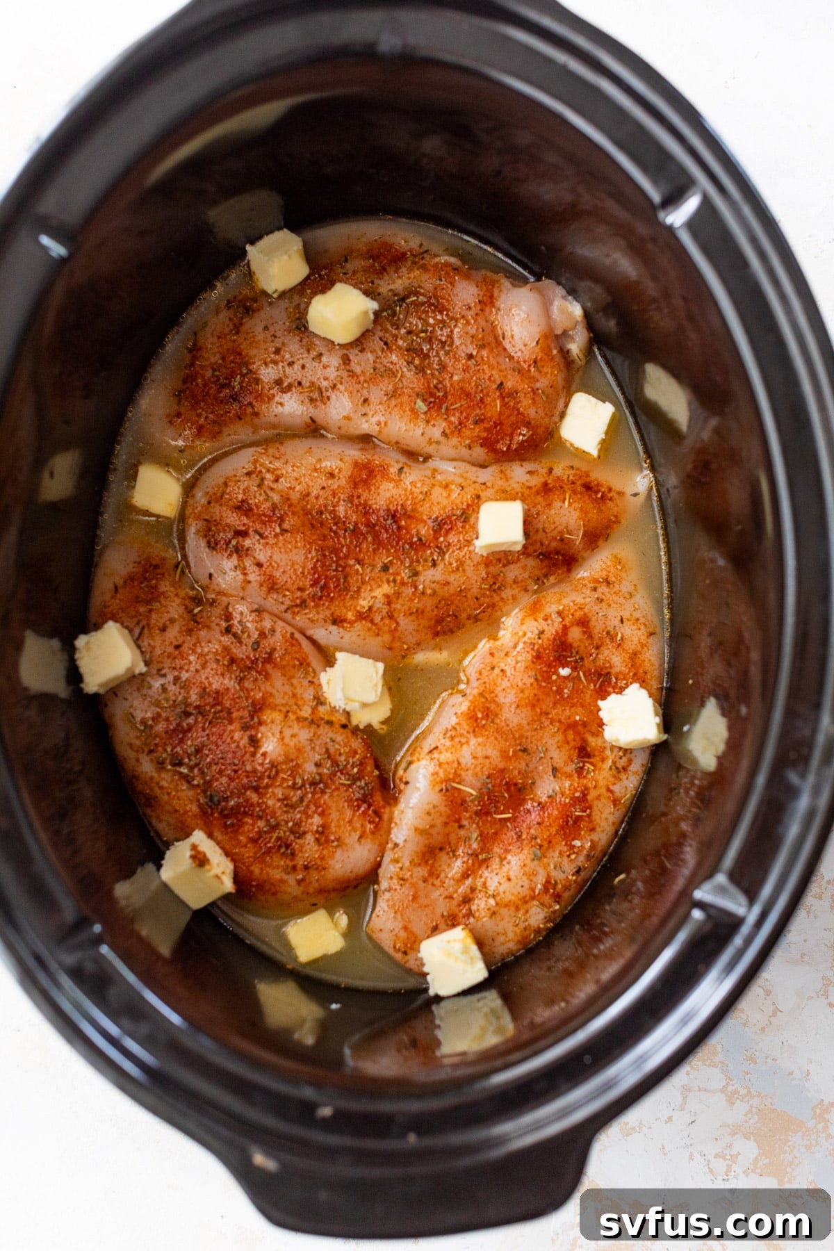 raw seasoned chicken breasts in a slow cooker with broth and pats of butter.